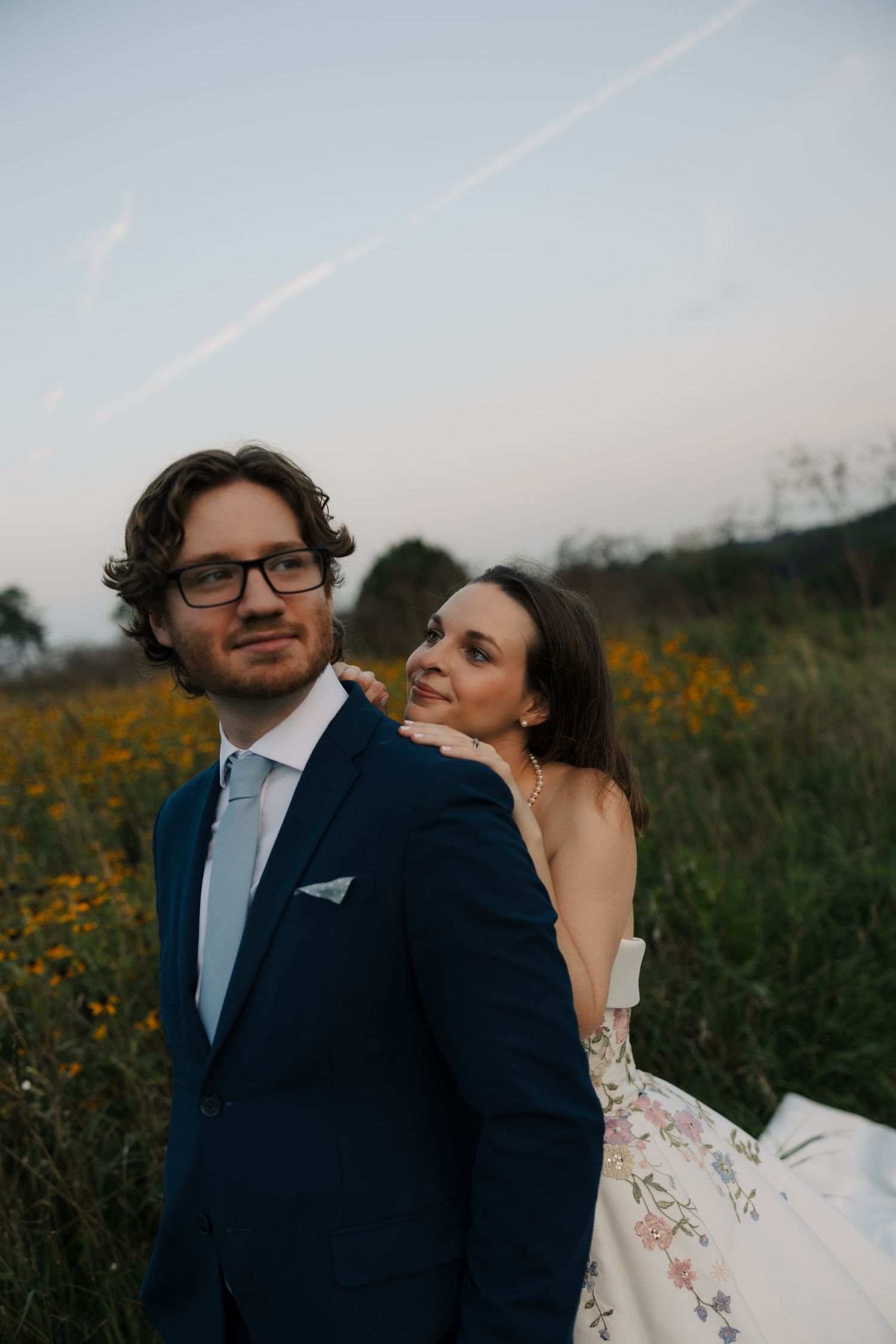 Bride in floral wedding gown rests chin on groom's shoulder as he gazes forward in a wildflower field.