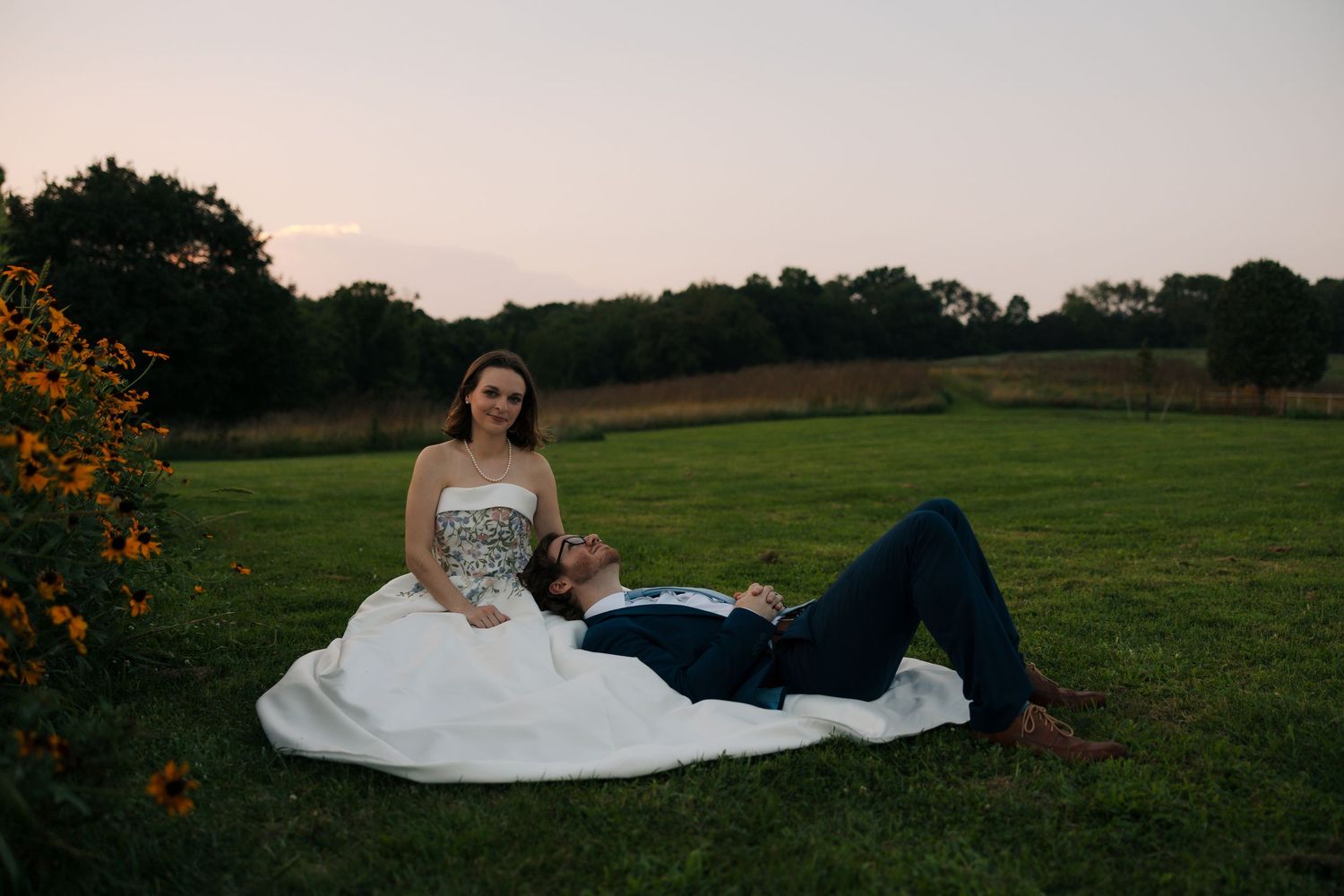 Bride and groom relaxing on green lawn at sunset, groom lying across bride's lap near orange flowers.