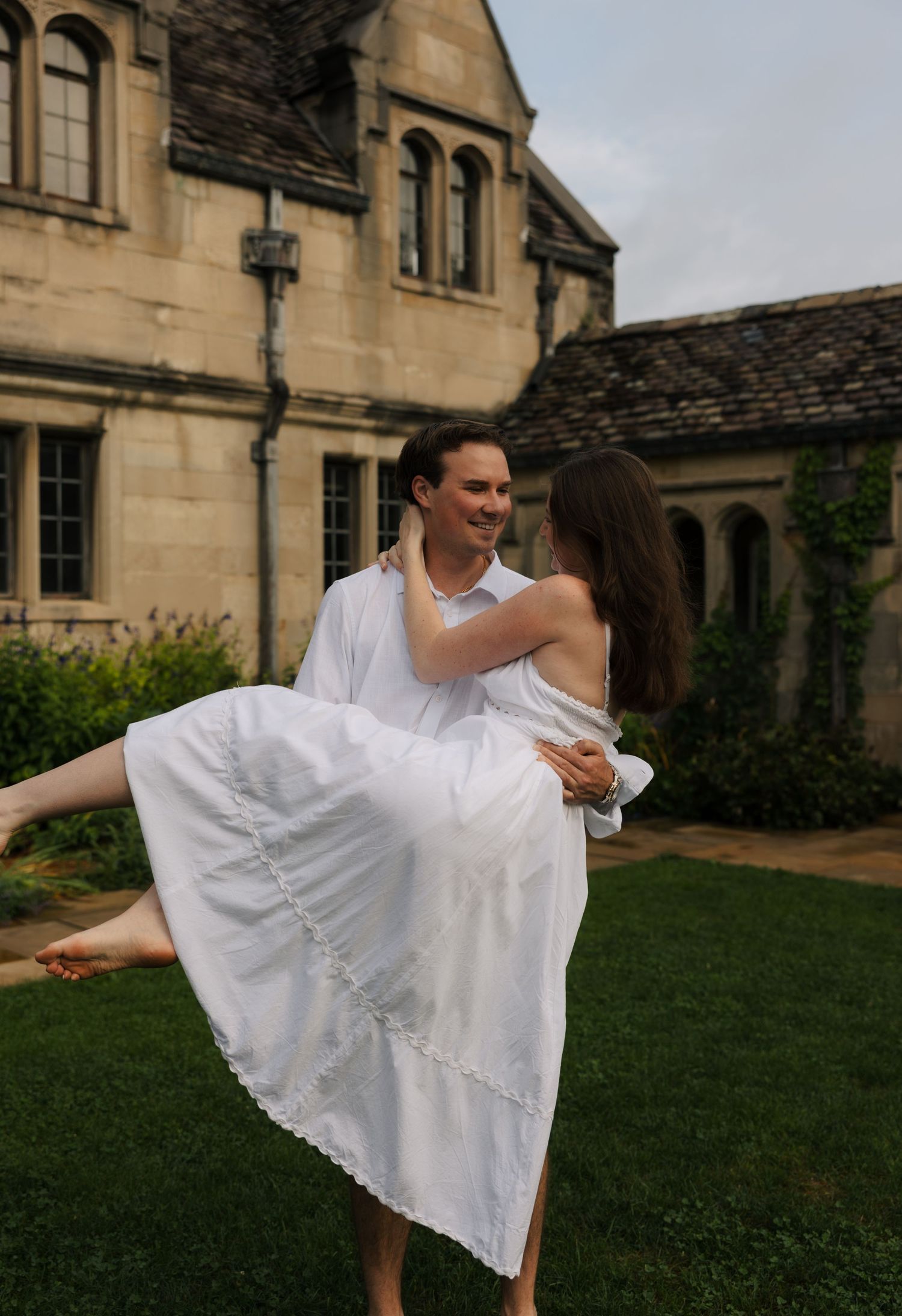 Man in white shirt carrying woman in white dress outdoors near stone manor house on green lawn.