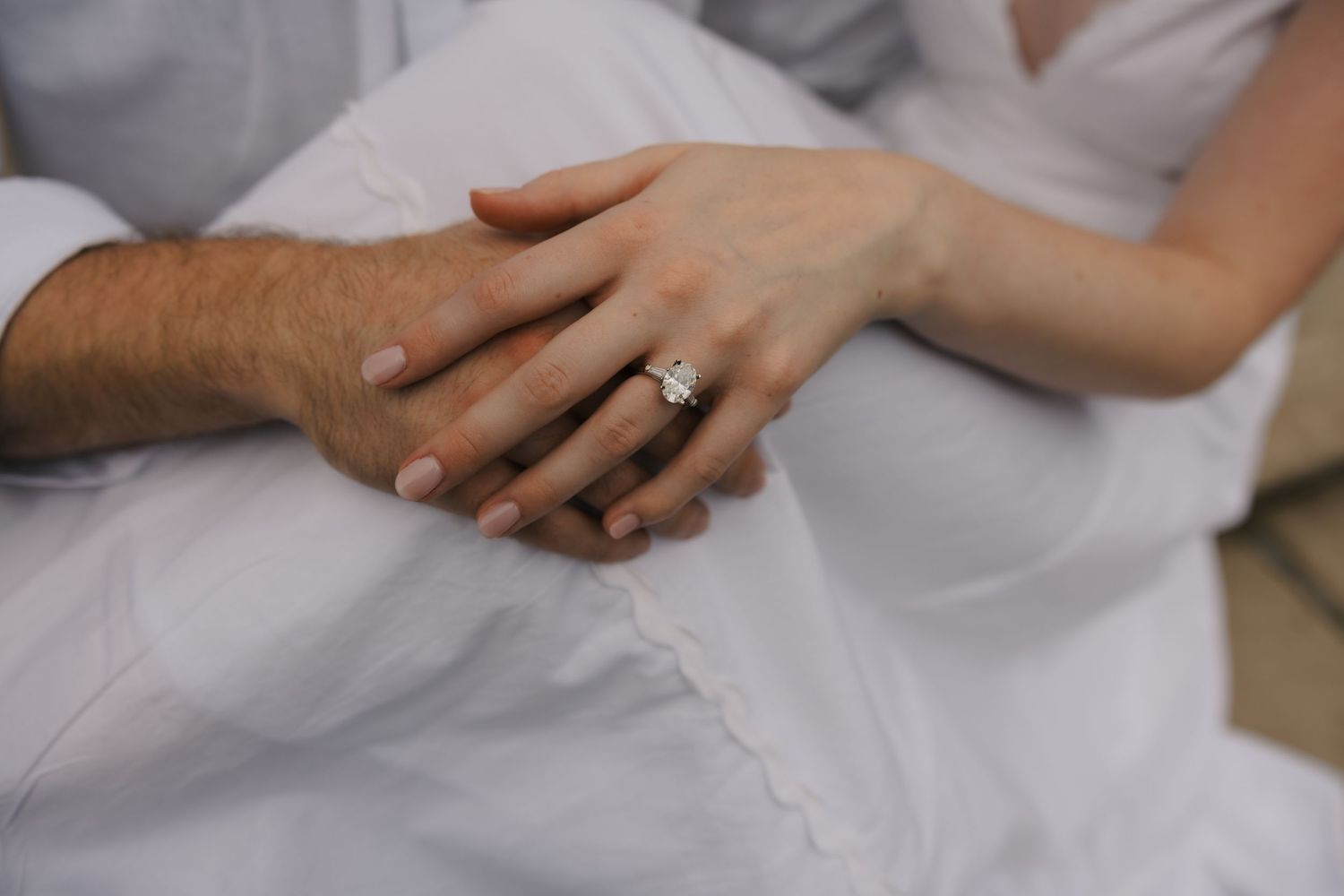 Close-up of a woman's hand with a diamond engagement ring resting on a white dress.