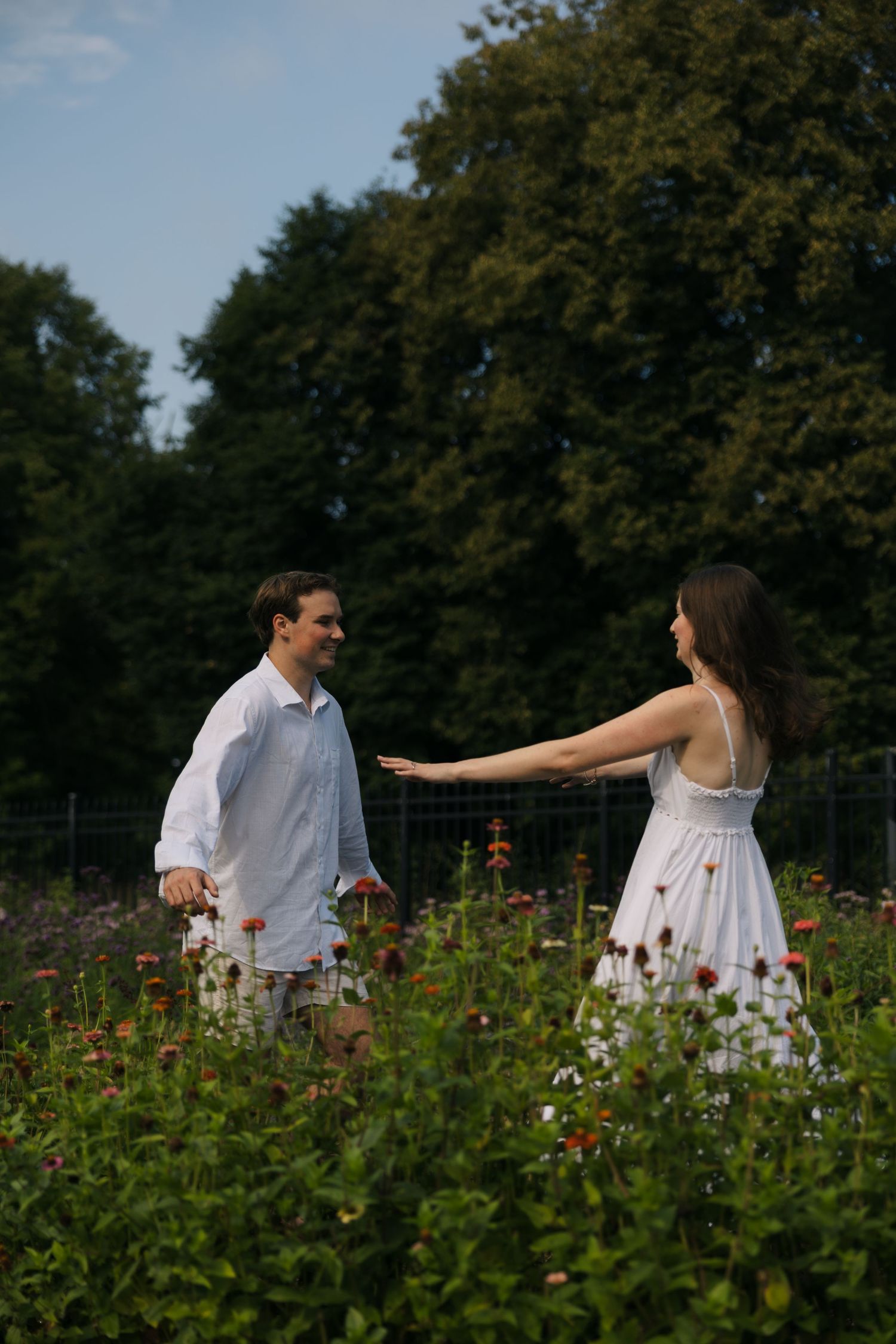 Couple reaching for each other's hands while walking through a vibrant flower garden with tall trees.