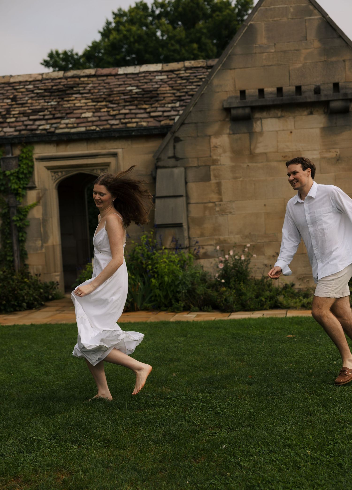 Joyful woman in white dress runs barefoot across green lawn with smiling man behind her.