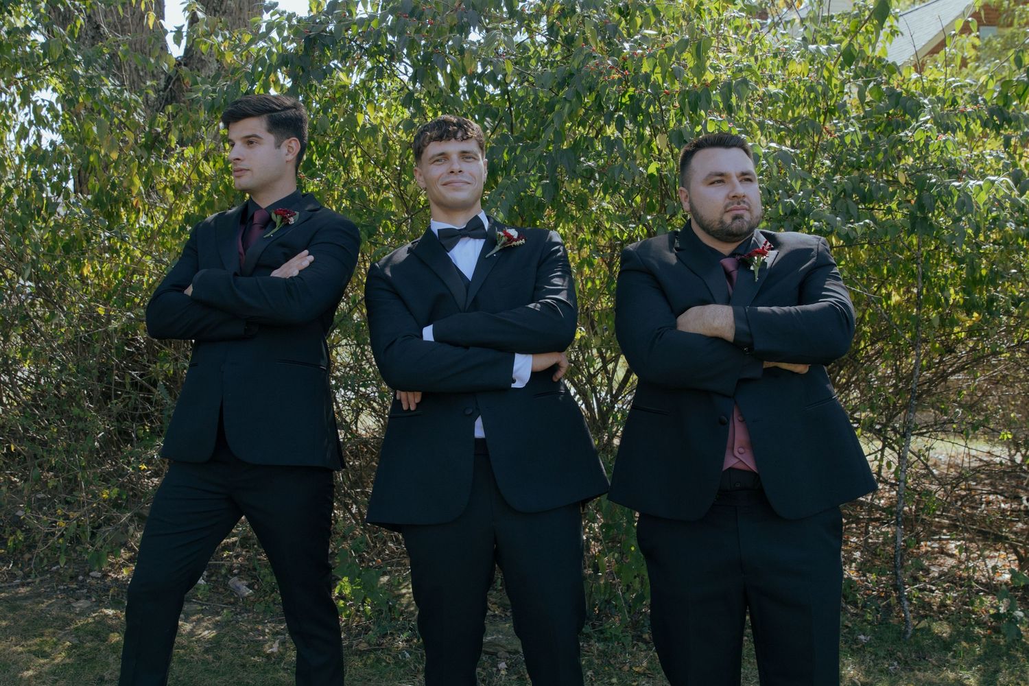 Three men in black suits stand with arms crossed in front of green foliage outdoors.