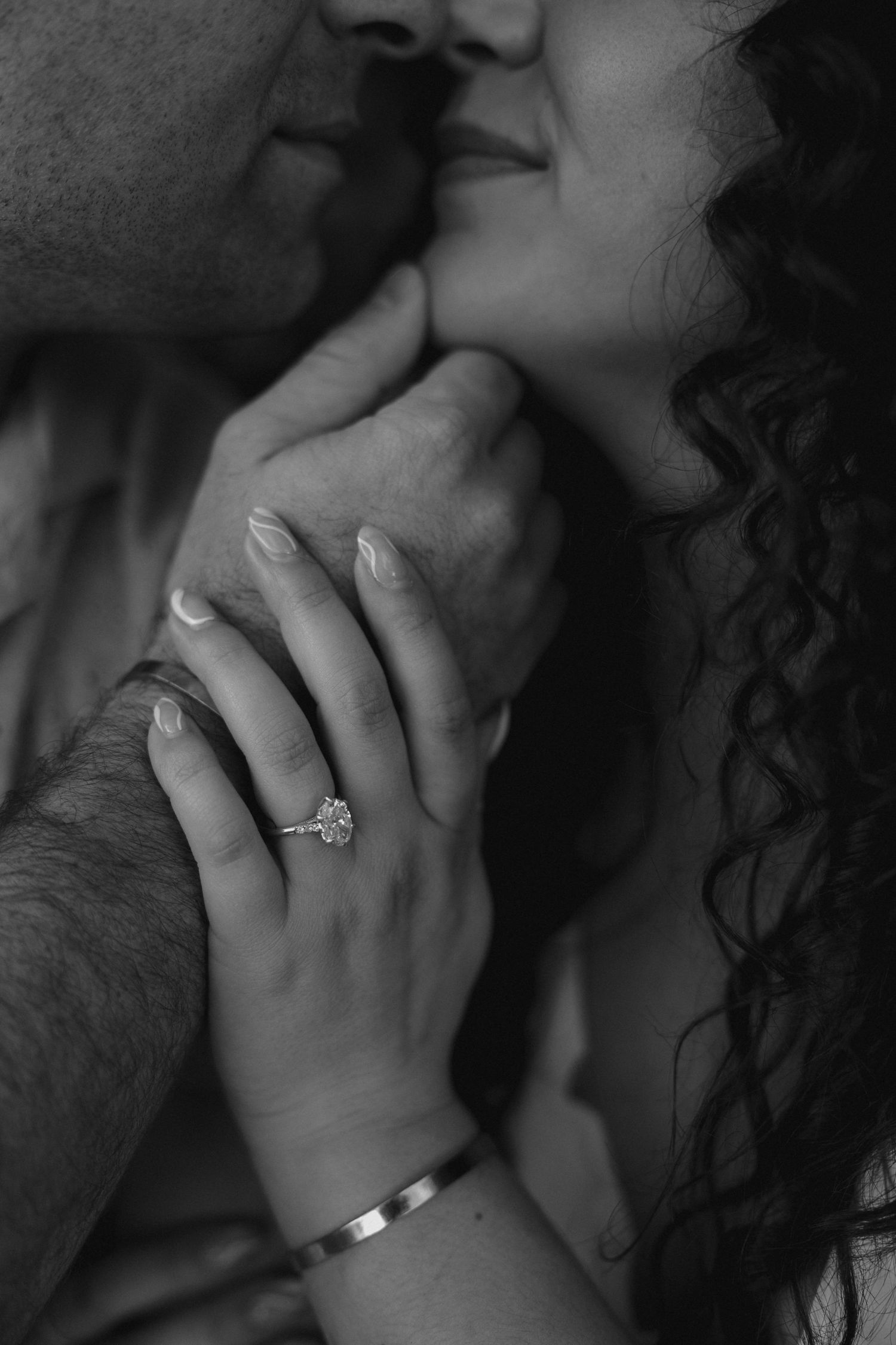 Black and white close-up of engaged couple almost kissing, showcasing diamond engagement ring and bracelet.