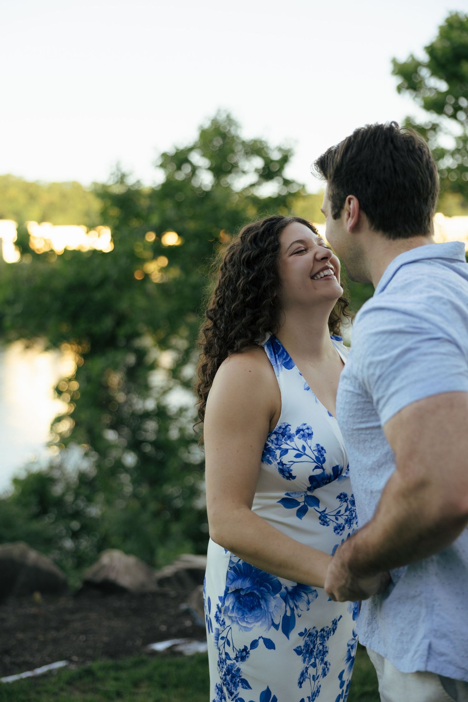 Happy couple holding hands and laughing together near a serene lakeside at golden hour.
