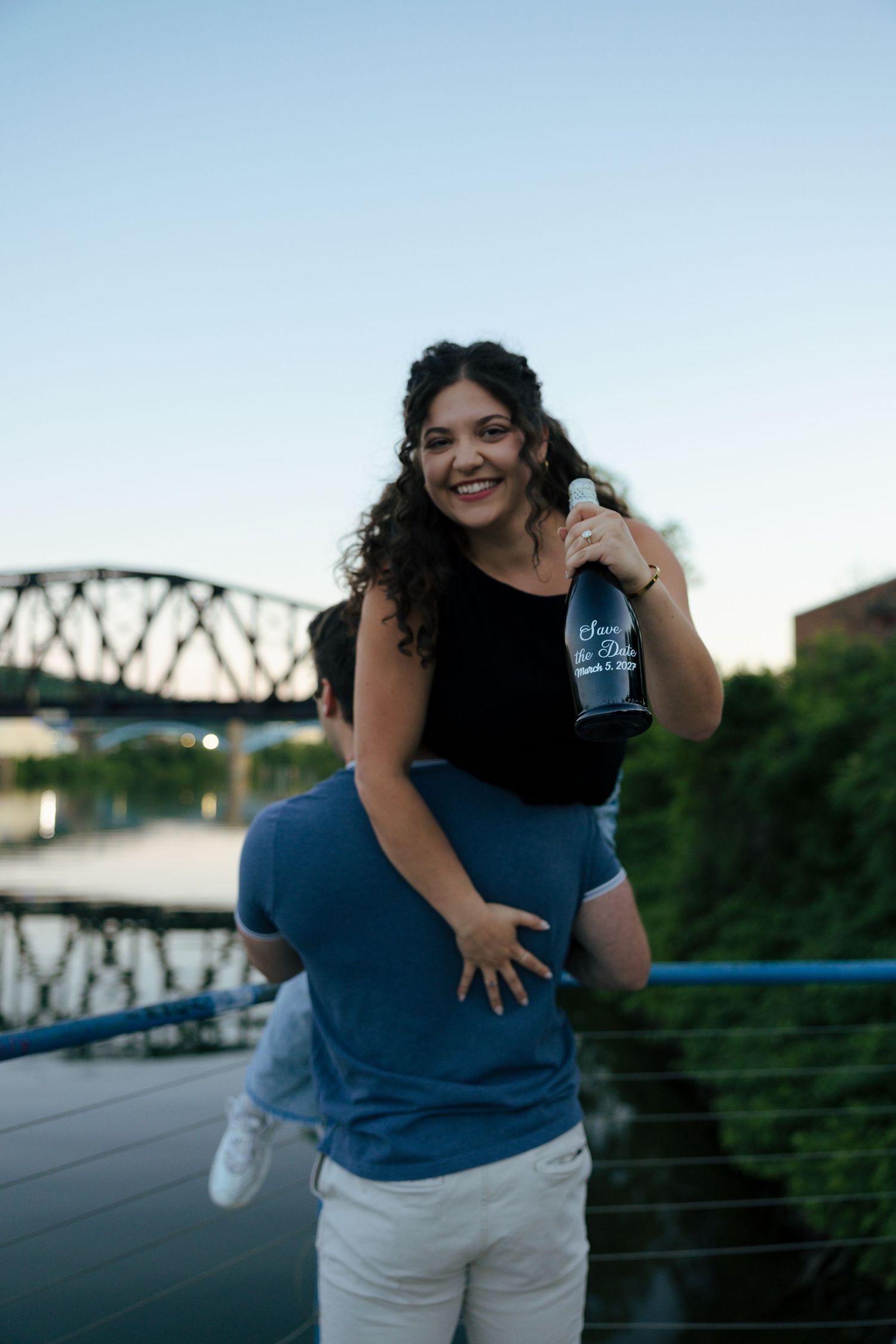 Woman laughing while being carried piggyback by man on bridge, holding champagne bottle at sunset.