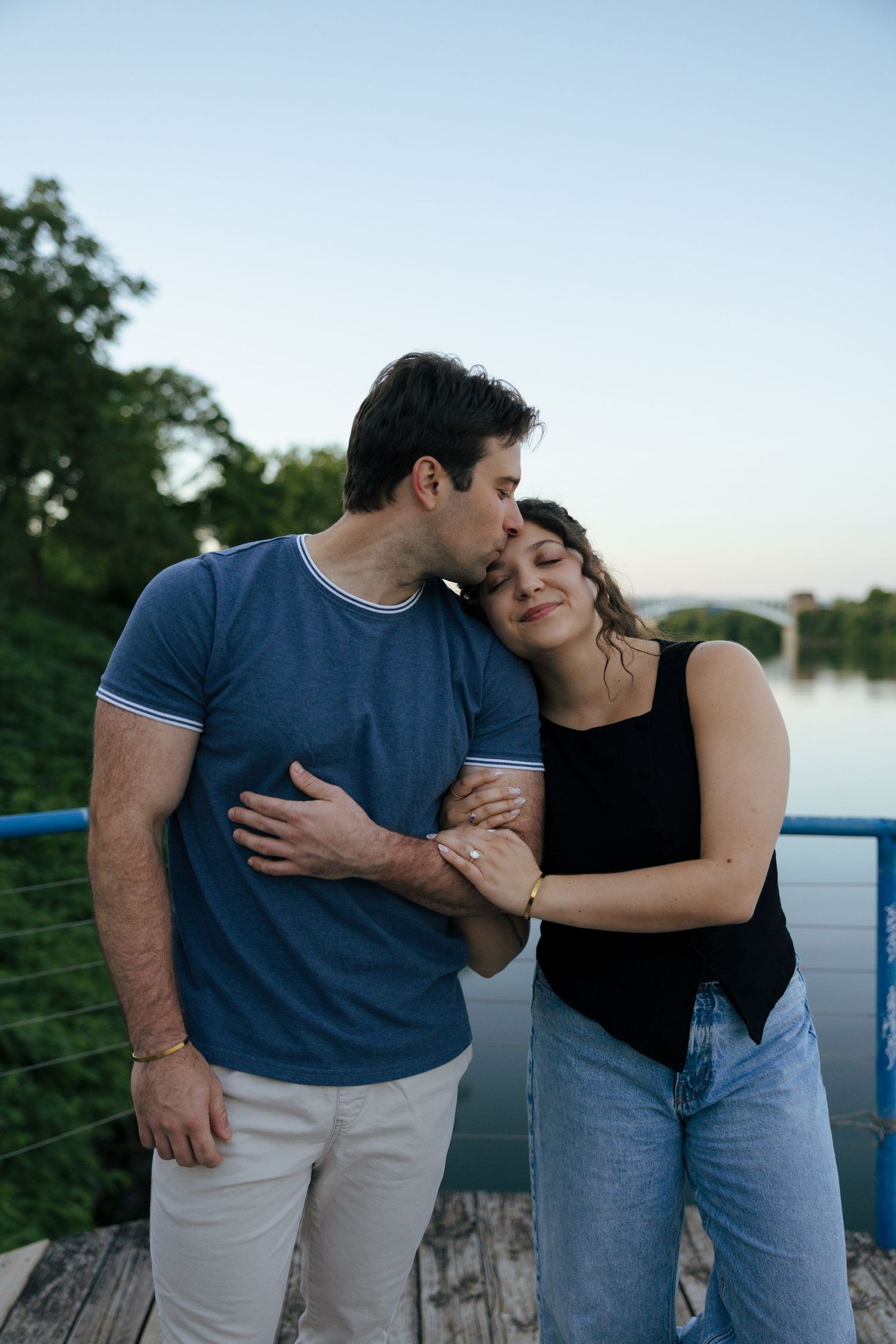 A man in a blue shirt kisses a woman's head as they embrace on a waterfront bridge at sunset.