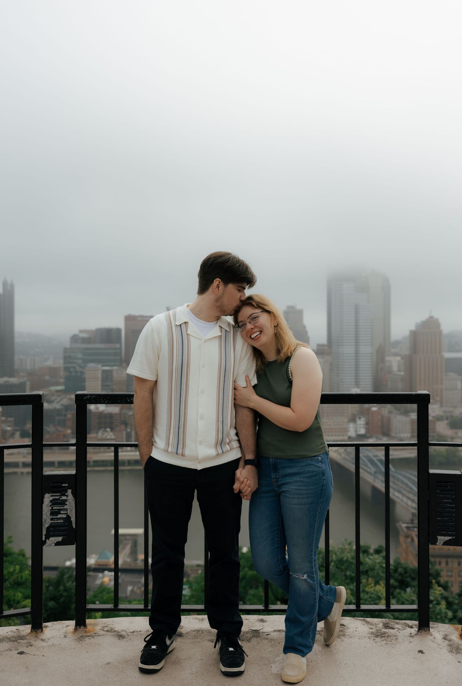 Happy couple holding hands and smiling on a rooftop overlook with Pittsburgh skyline in the misty background.