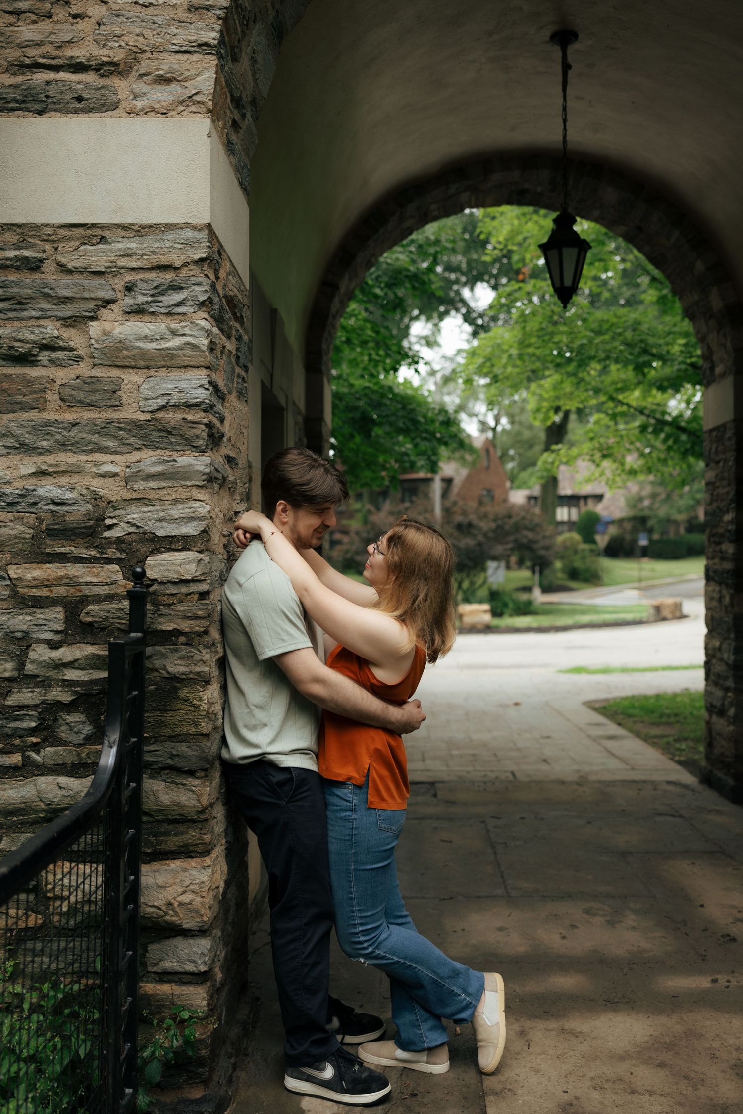 Couple embracing against a stone archway pillar in a charming outdoor corridor surrounded by lush greenery.