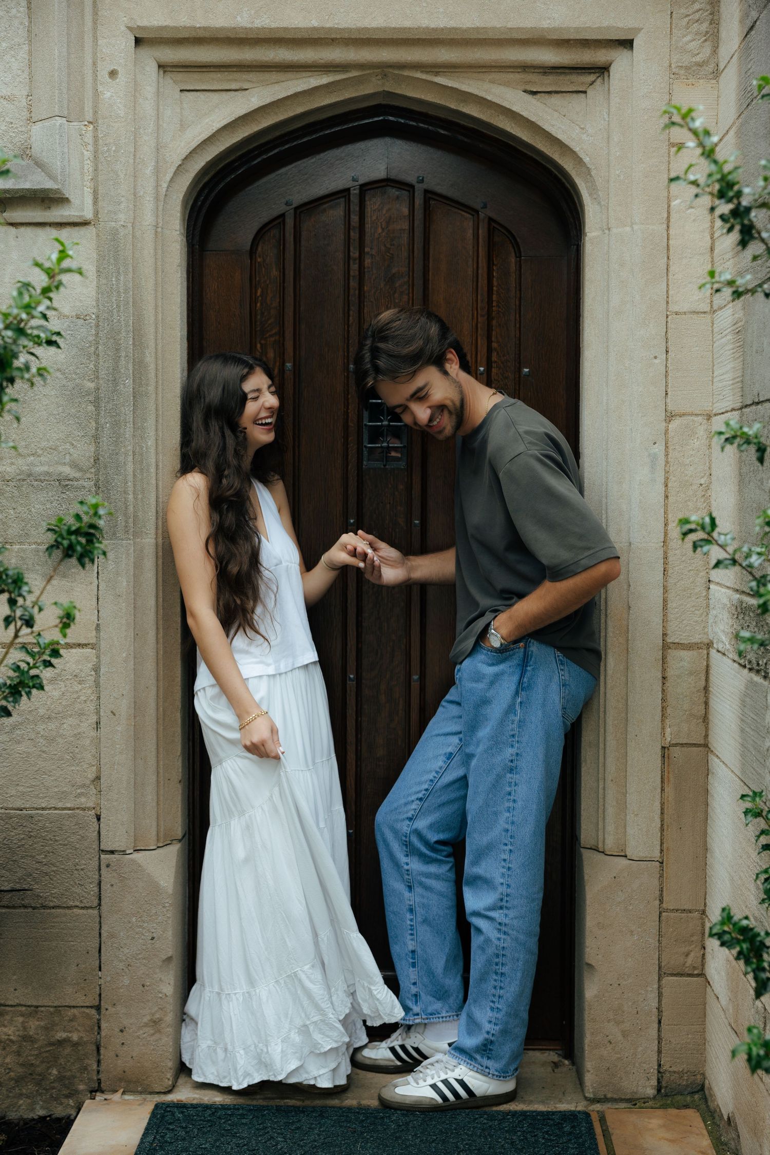 Happy couple laughing and holding hands in front of ornate arched wooden door of stone building.