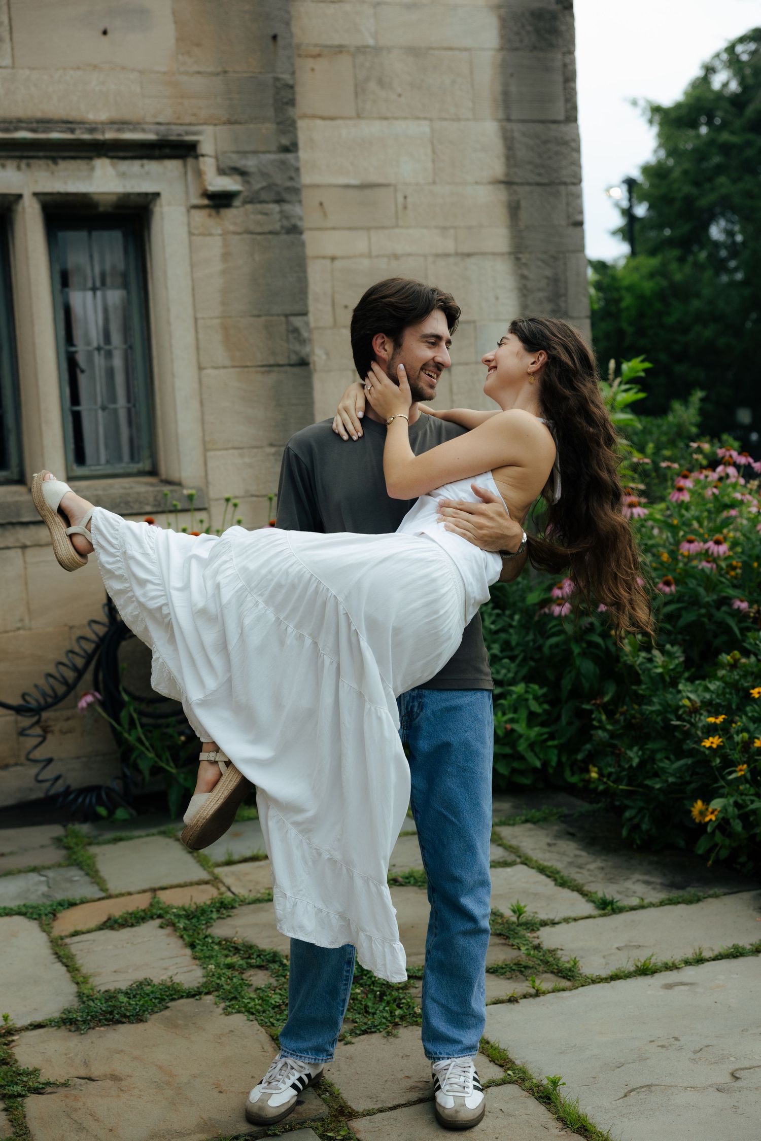 A joyful couple laughs as the man carries the woman in a white dress outdoors.
