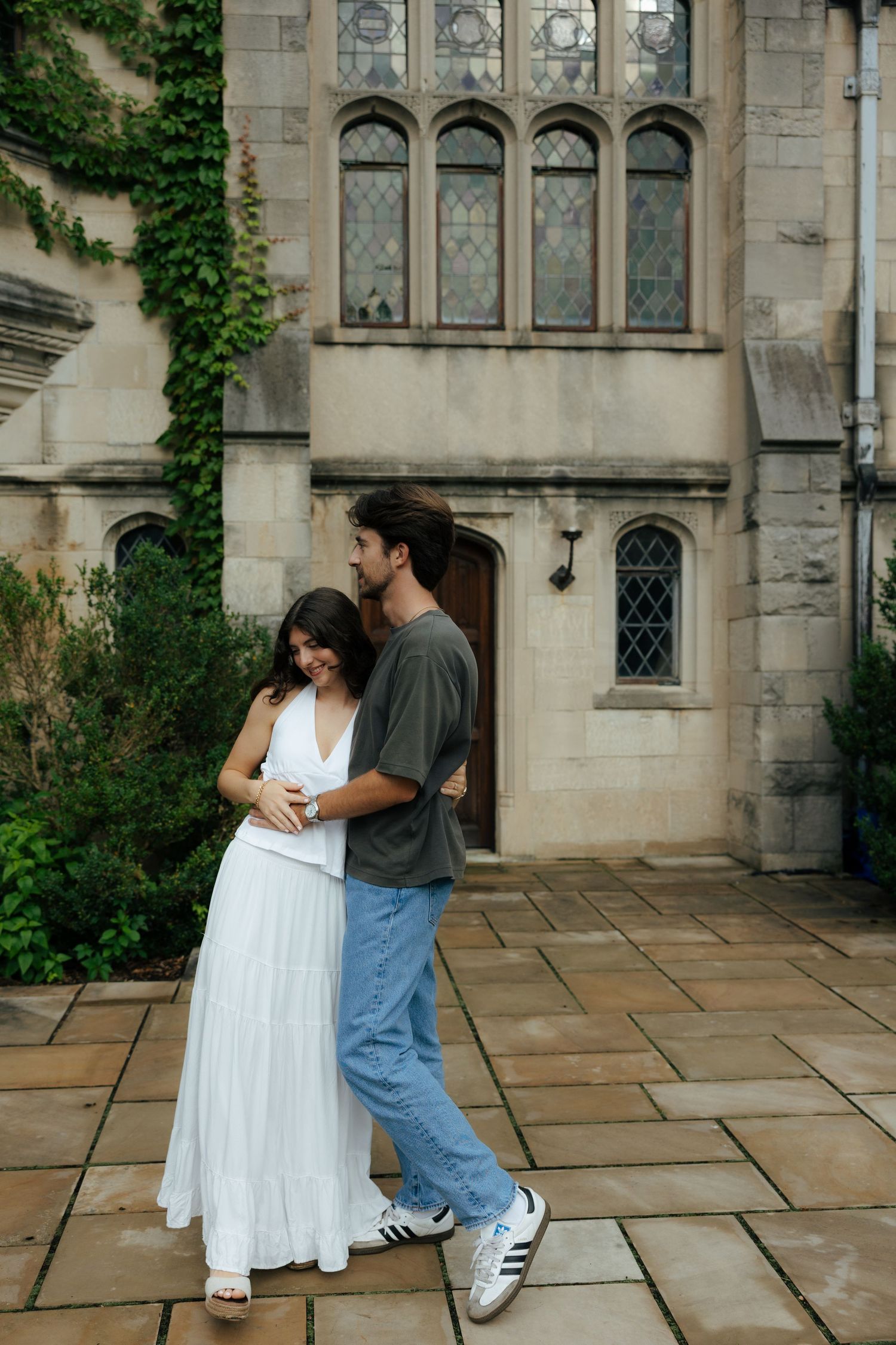 Couple embracing and smiling outside a gothic stone building covered in ivy on a cloudy day.
