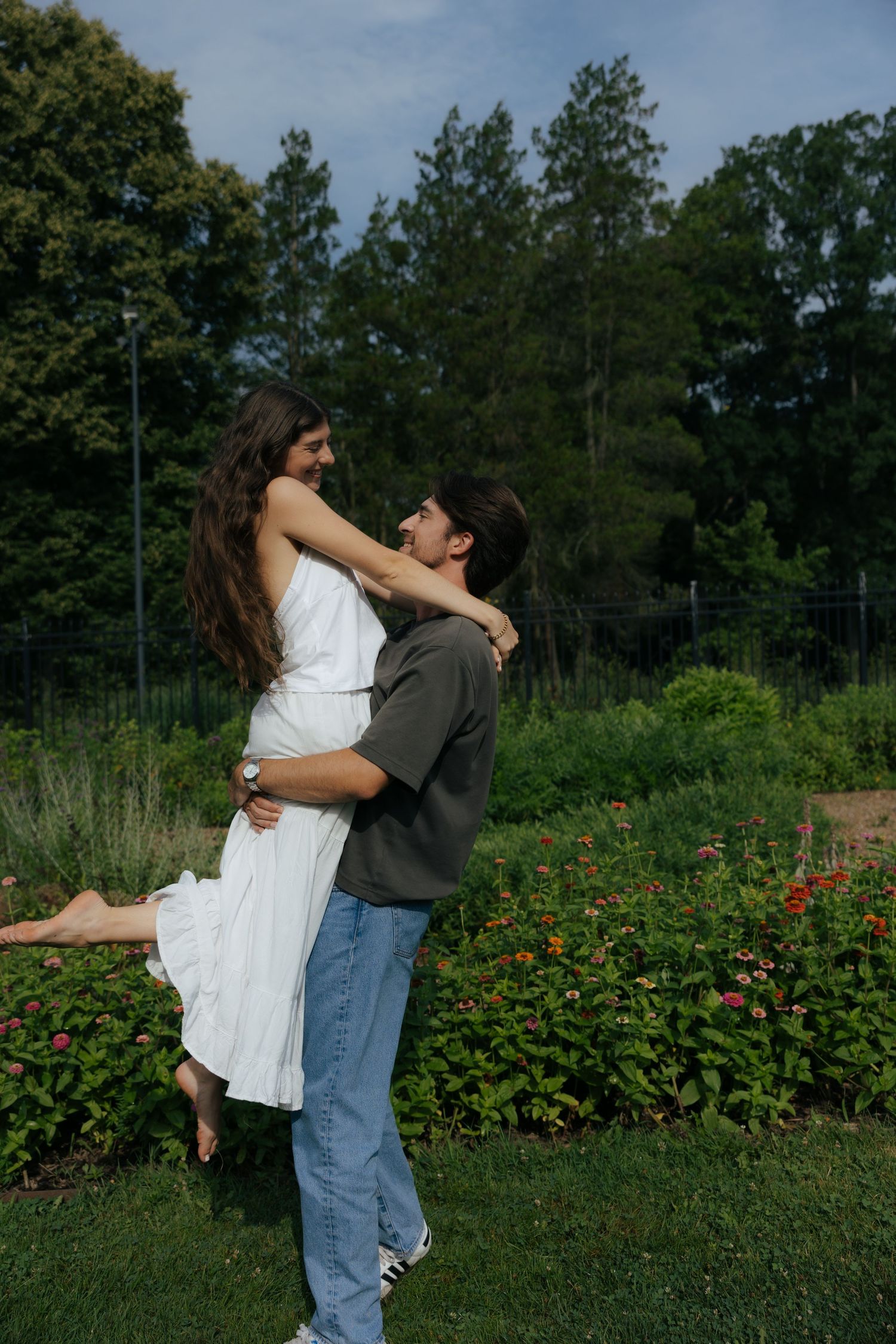 A couple shares a joyful moment as the man lifts the woman in a white dress in a sunny garden setting.