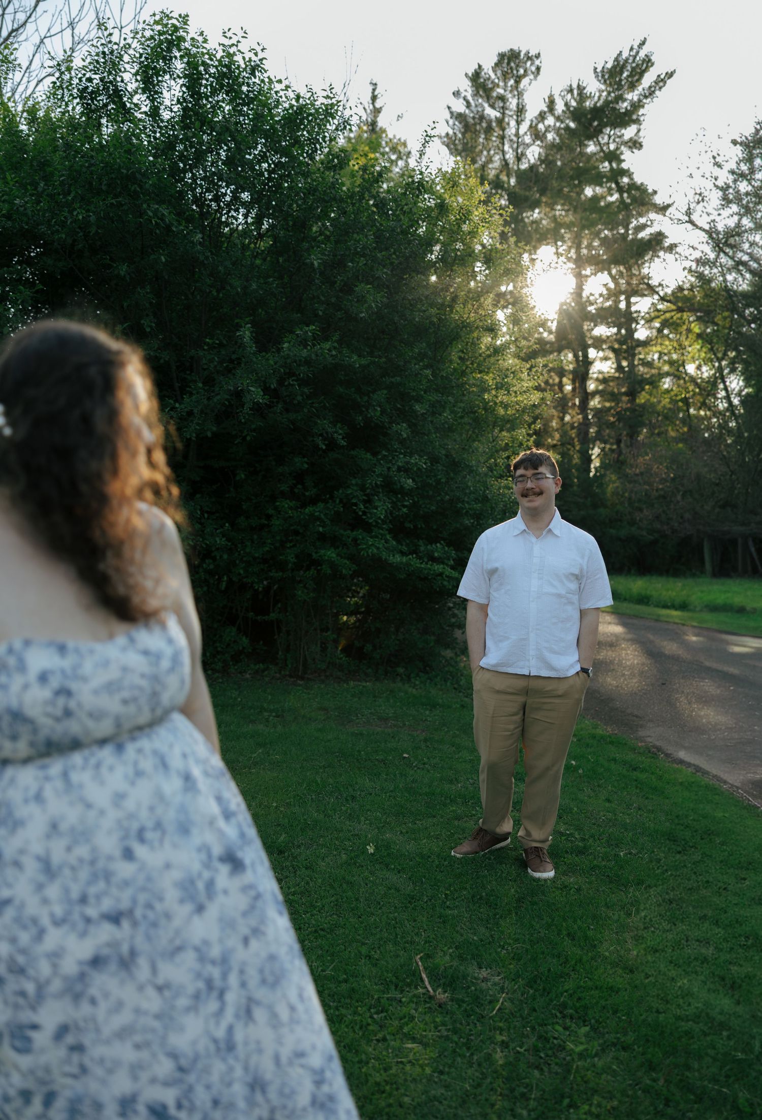 Couple standing apart in a sunlit garden park, man in white shirt and khakis smiling at woman in floral dress.