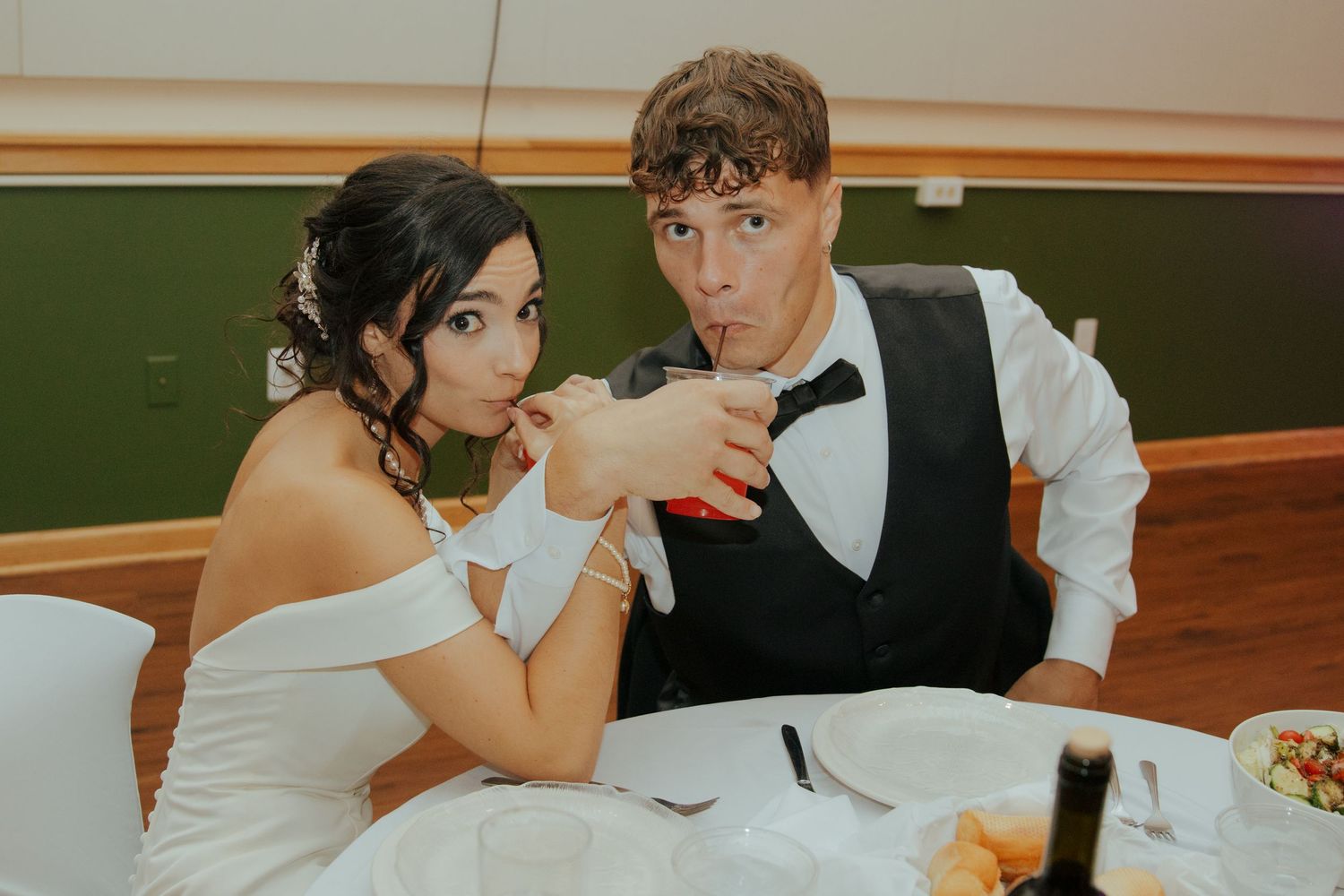 Bride and groom playfully sharing a drink together at wedding reception table.