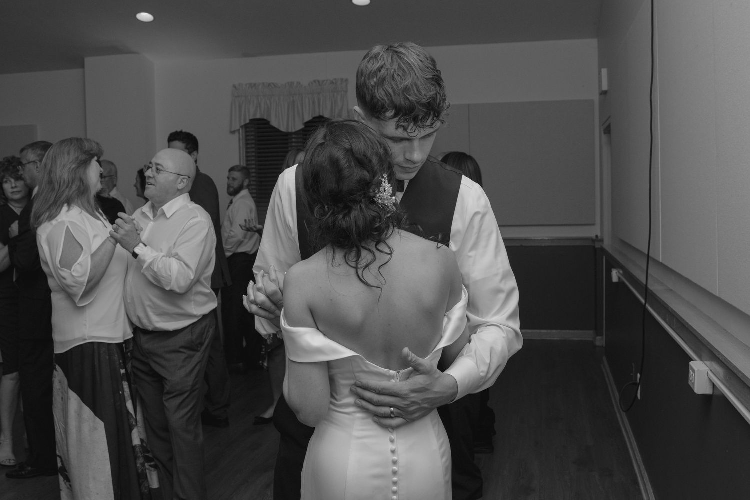 Bride and groom sharing first dance while guests dance in background at wedding reception in black and white.