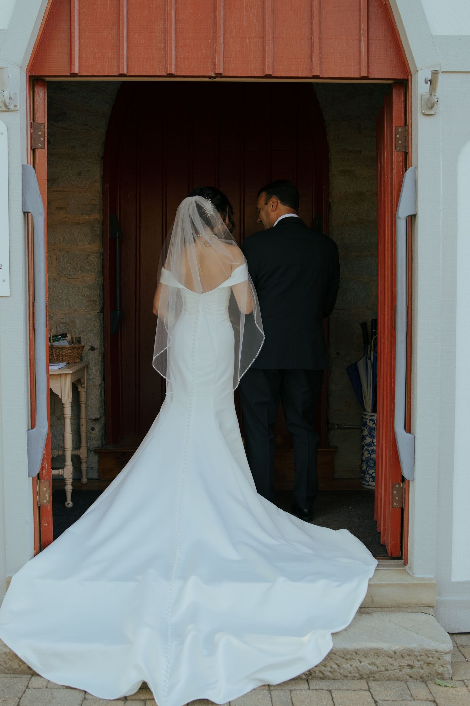 Bride in white mermaid gown with veil and groom entering church doors before ceremony.
