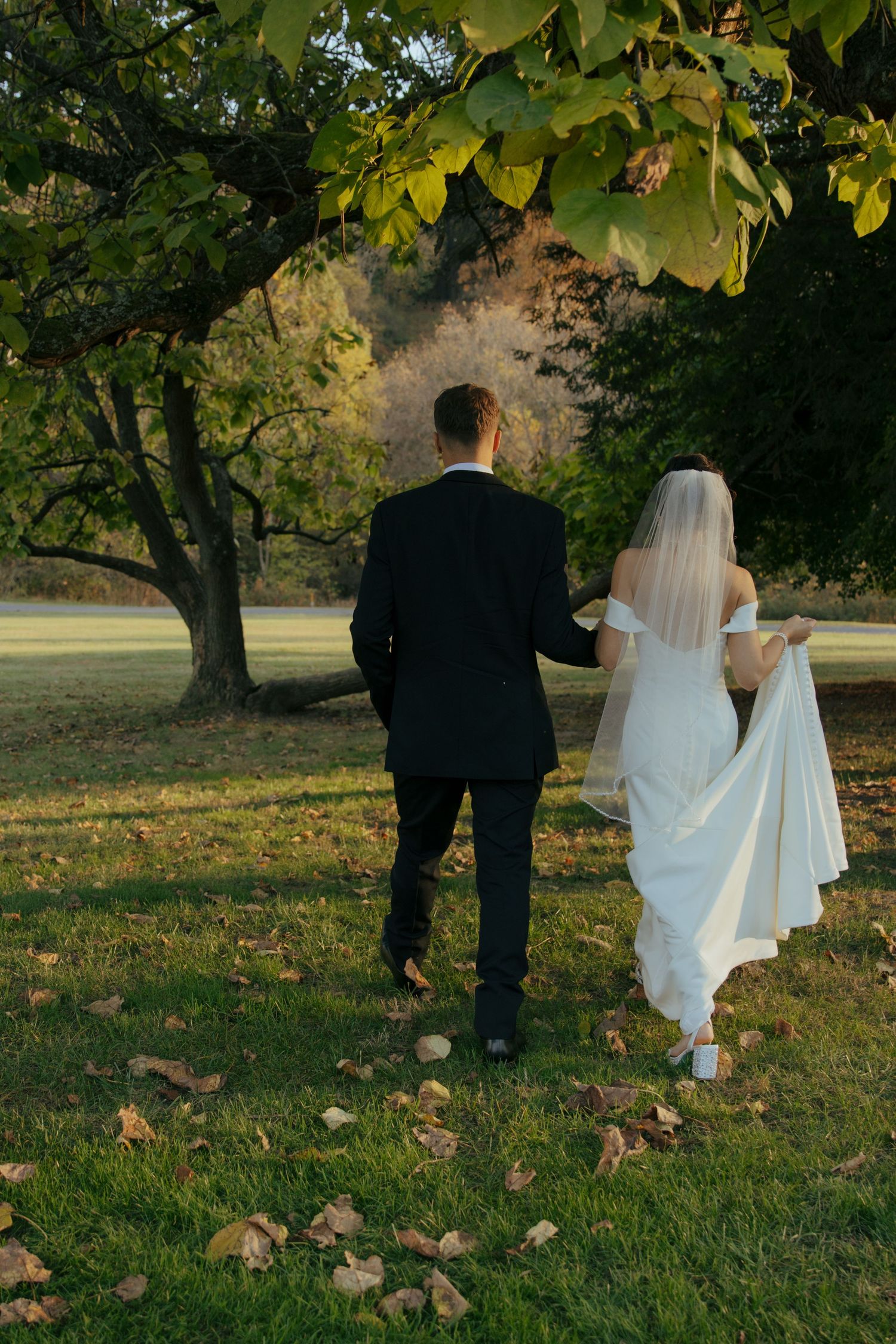 Bride and groom walking hand in hand through a lush green park with autumn leaves on the ground.