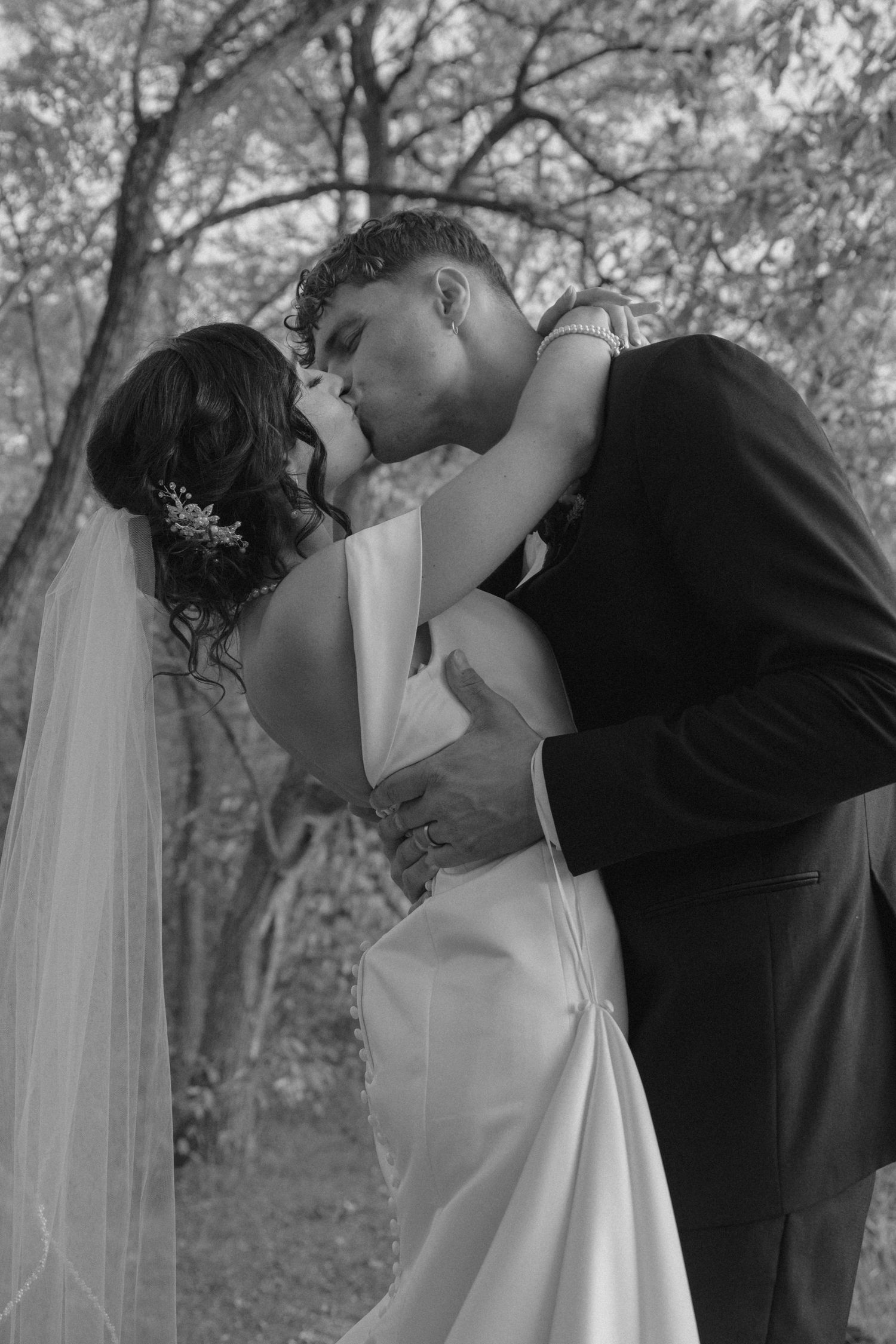 Groom dipping and kissing bride in a romantic black and white outdoor wedding portrait among bare trees.