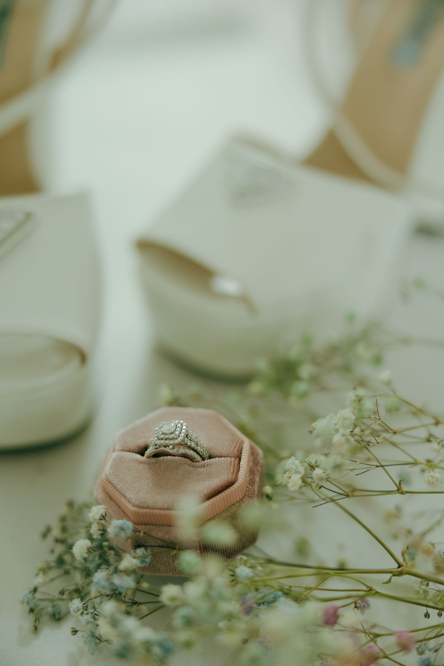 A diamond halo engagement ring displayed in a pink velvet box surrounded by baby's breath flowers and white bridal heels.