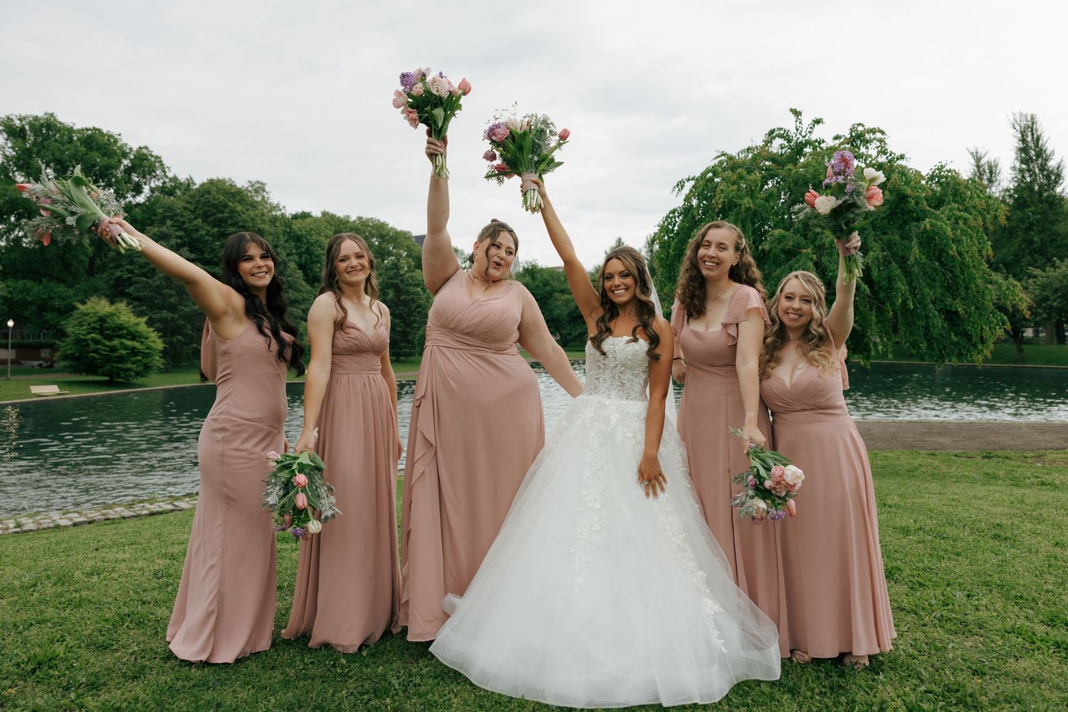 Bride in white gown poses with five bridesmaids in dusty rose dresses holding bouquets outdoors.