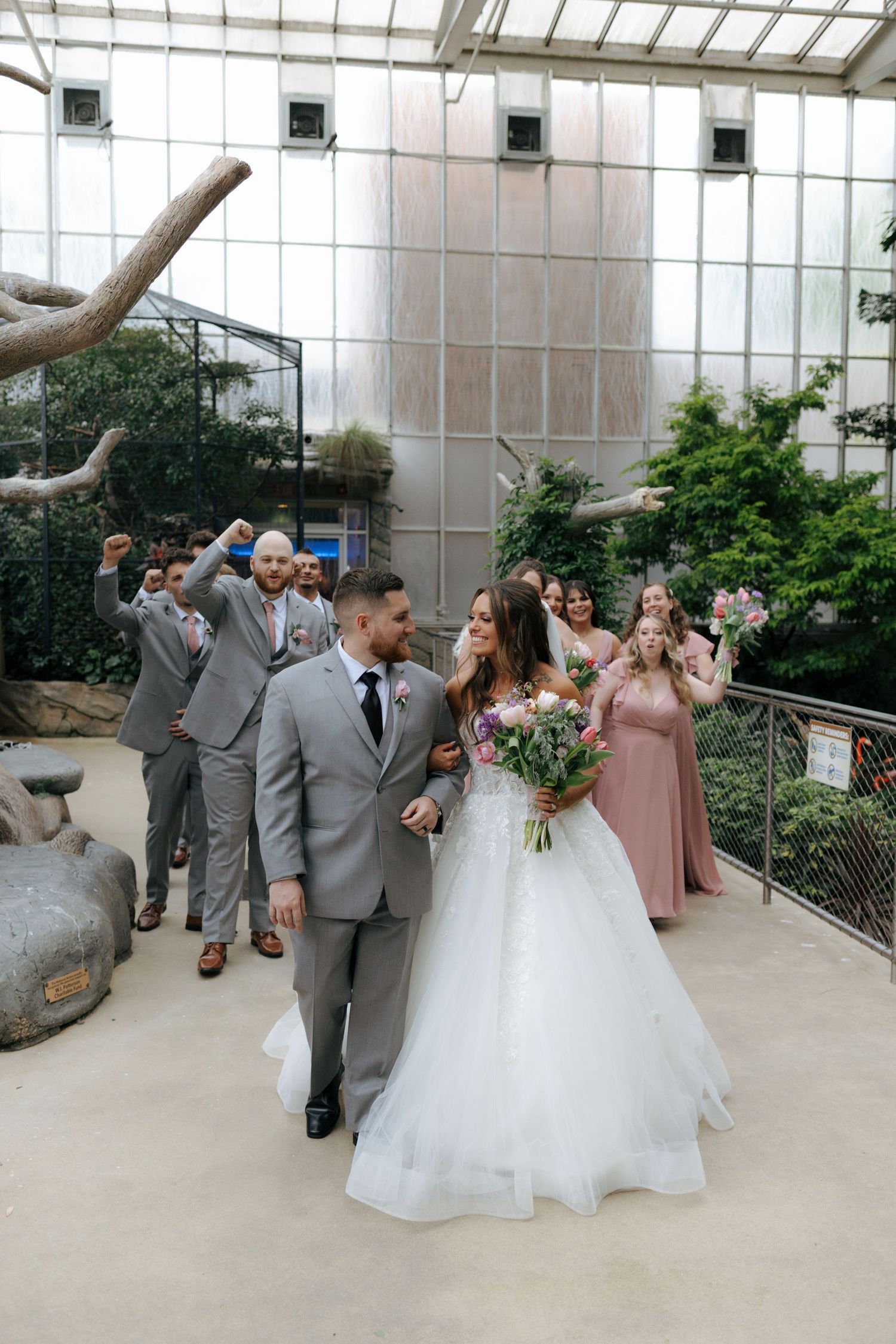 A bride and groom walk together as their wedding party cheers behind them inside a stunning greenhouse venue.