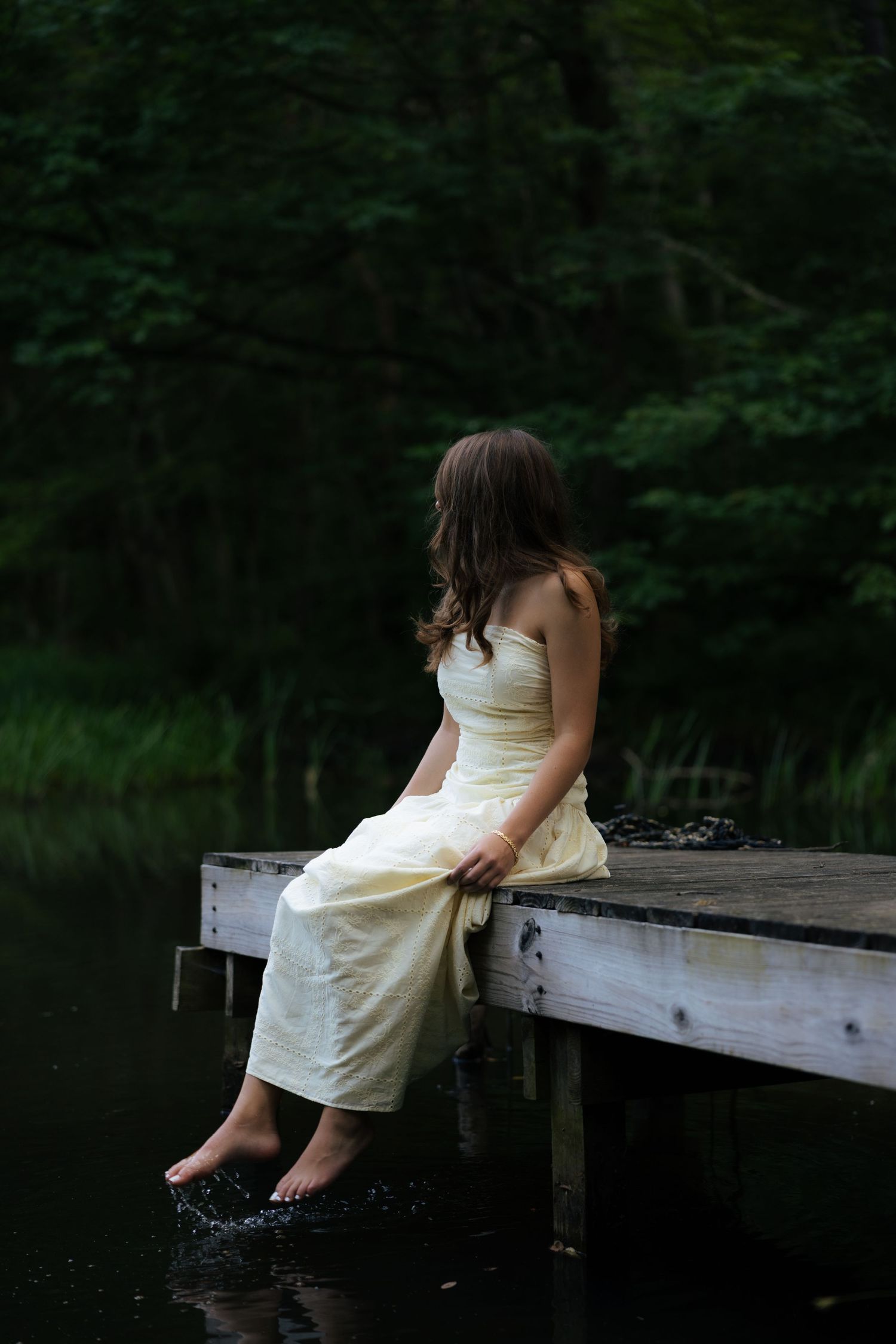 Woman in yellow dress sitting on wooden dock dangling feet in water by dark forest.