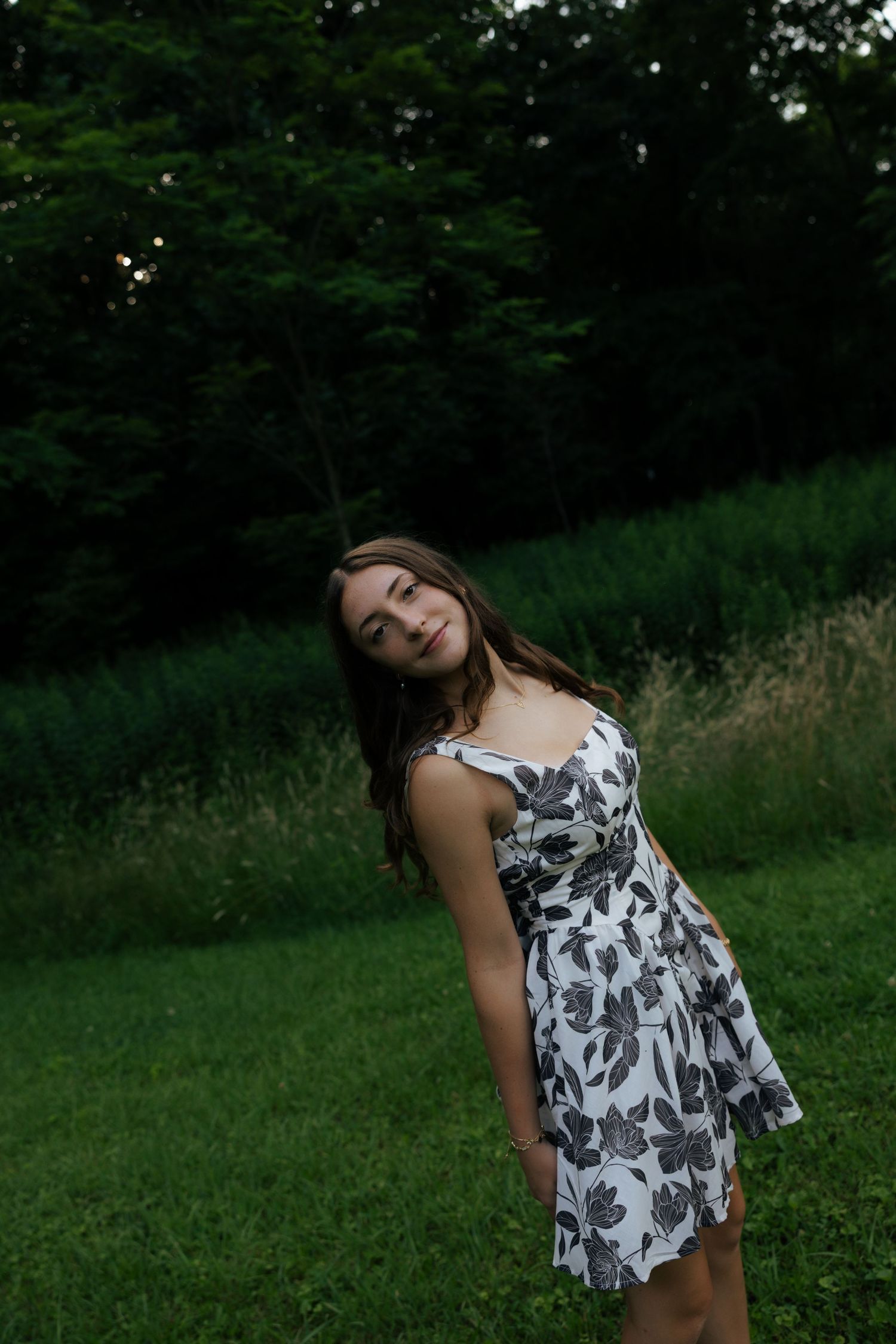 A teenage girl in a black and white floral dress poses on a grassy hill at dusk.