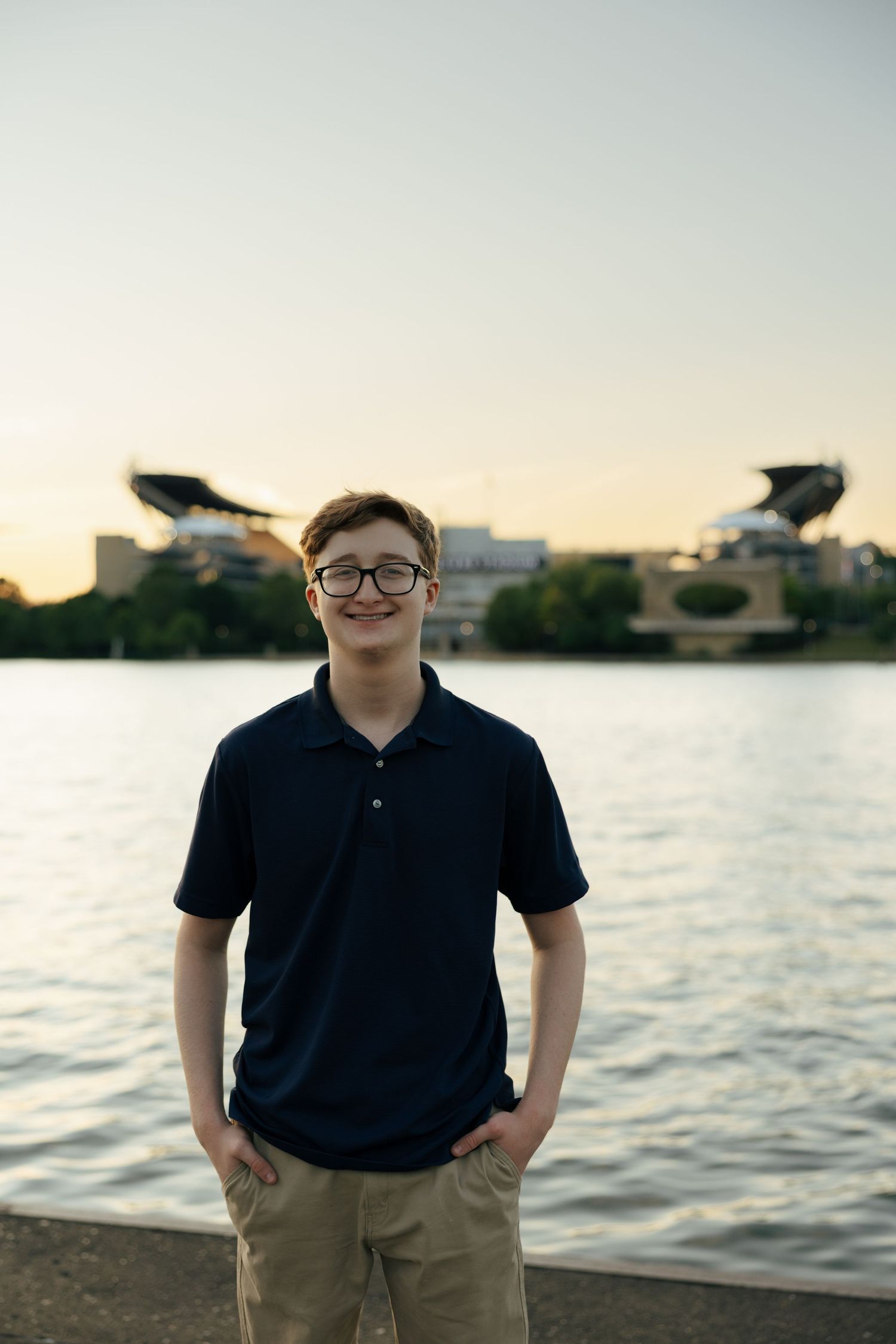 Smiling teen boy in navy polo stands by river at dusk with stadium in background.