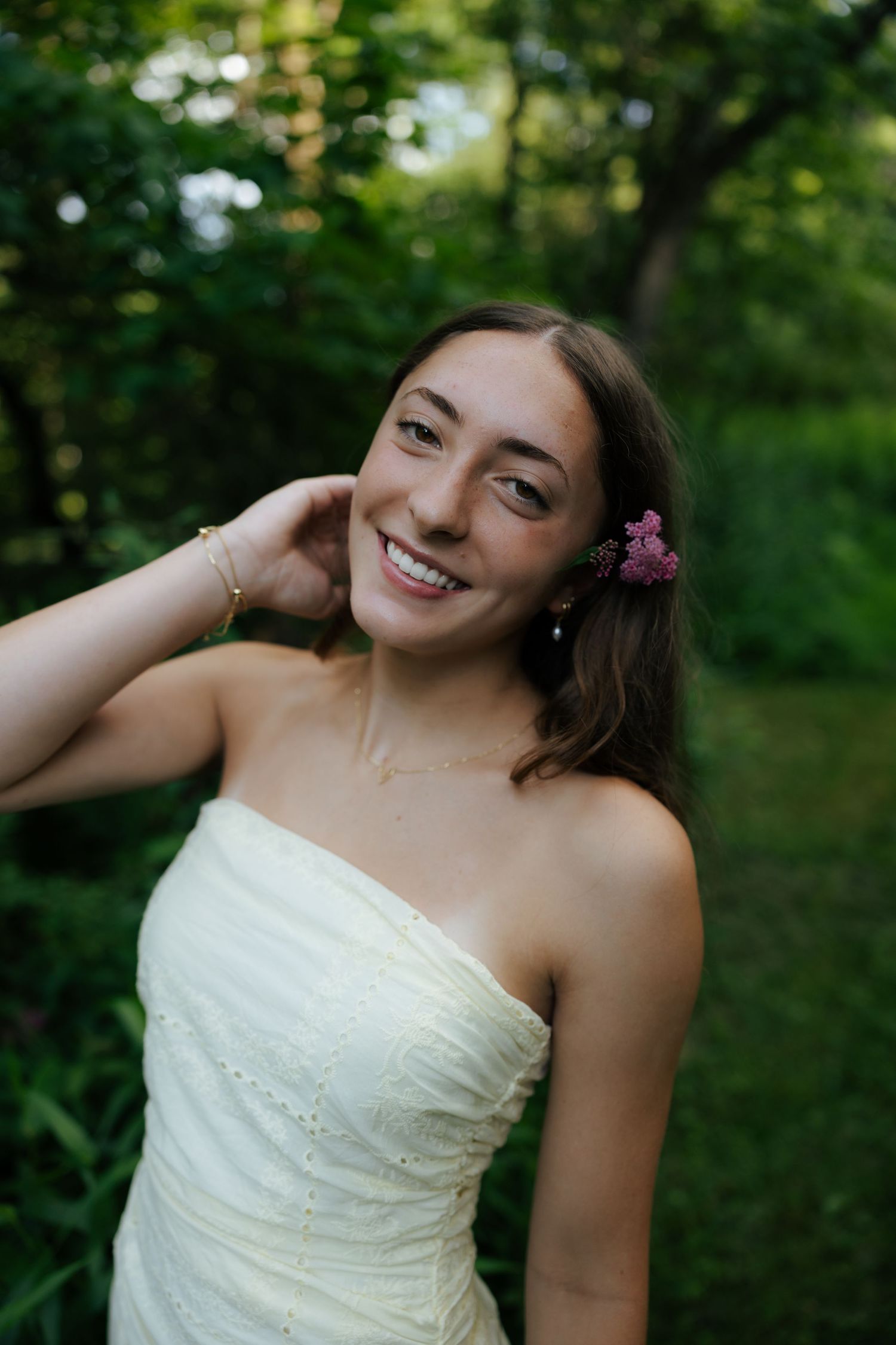 Smiling young woman with a pink flower in her hair wearing a strapless white dress outdoors.