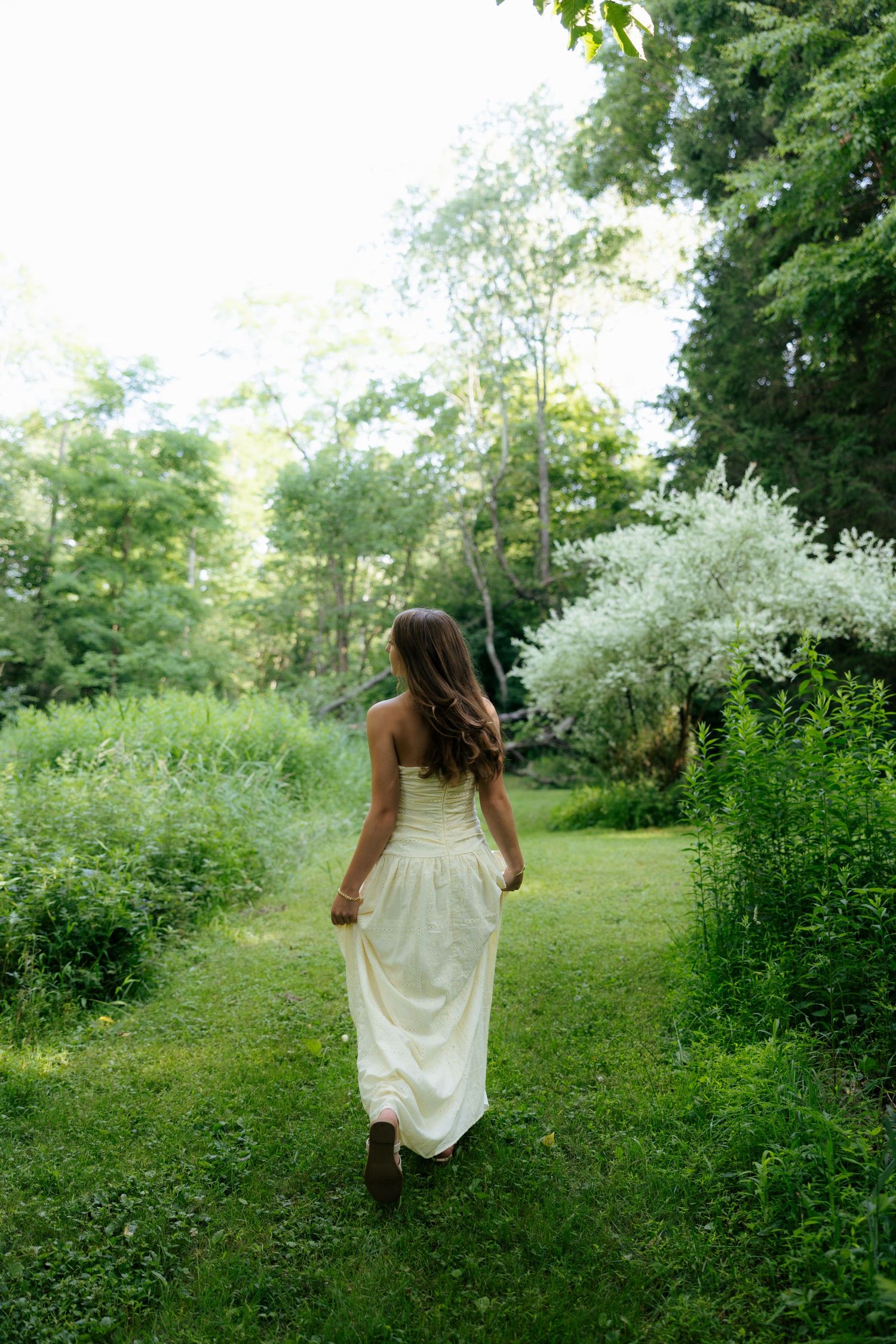 Young woman in white maxi dress walking away through lush green garden path toward blooming tree.