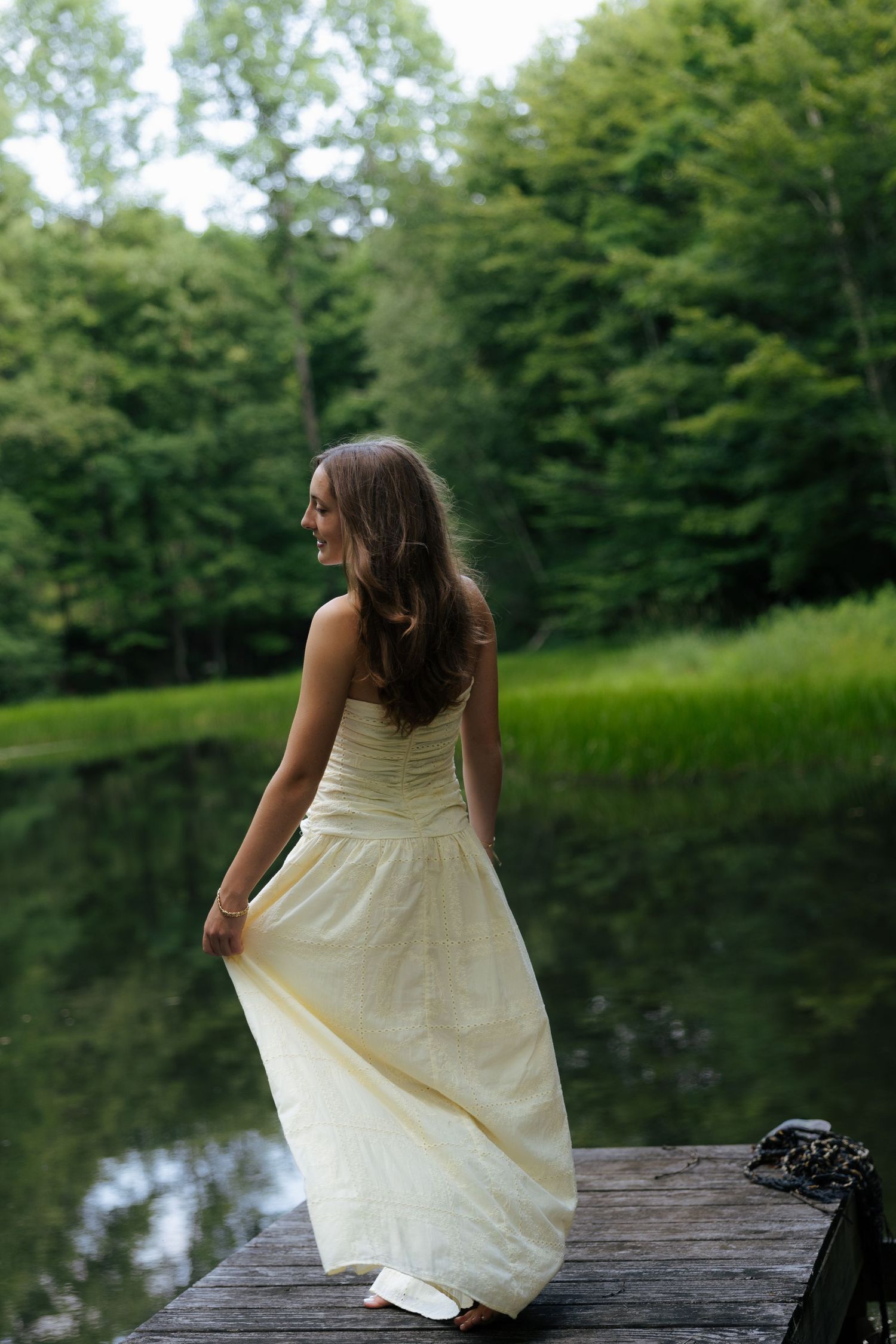 Woman in cream gown standing on wooden dock by serene lake.