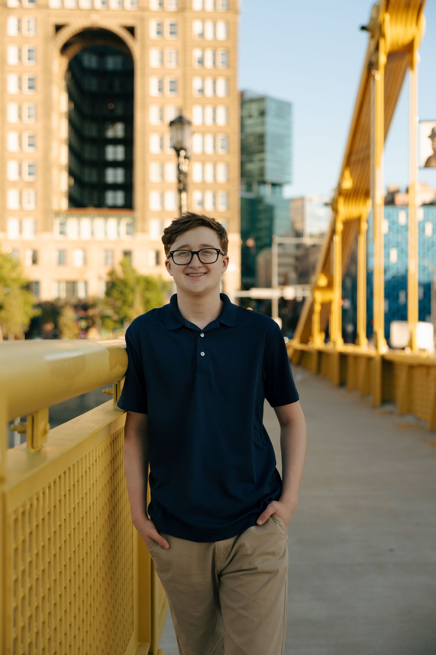 Teen boy in navy polo and glasses smiling on yellow Pittsburgh bridge with city buildings in background.