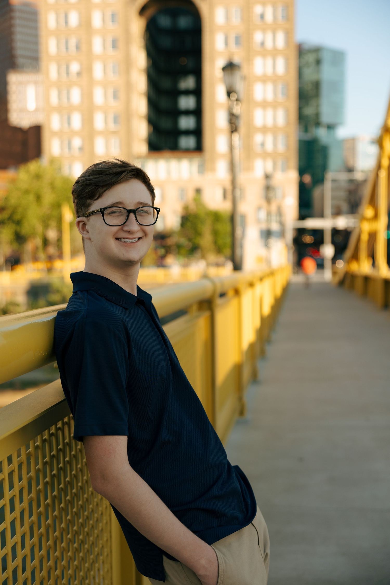 Smiling teenage boy with glasses in navy polo shirt posing on yellow pedestrian bridge in city.