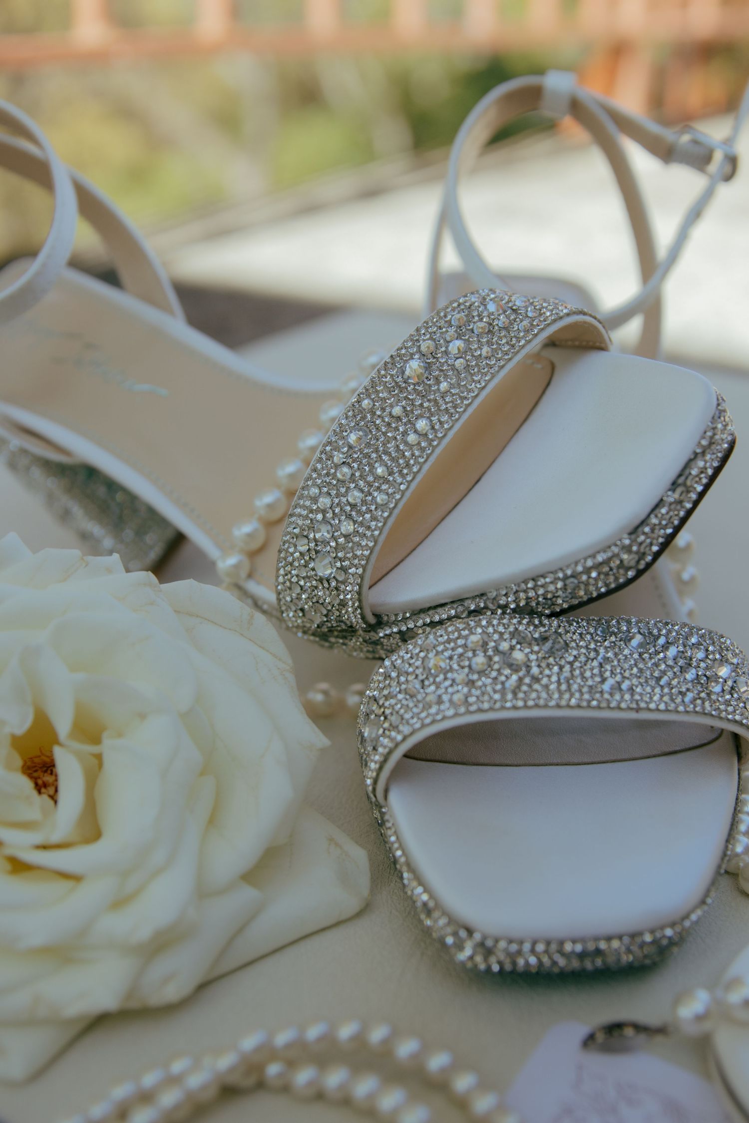 Crystal-embellished bridal heels with pearl details beside a white rose and pearl necklace on a wedding day.