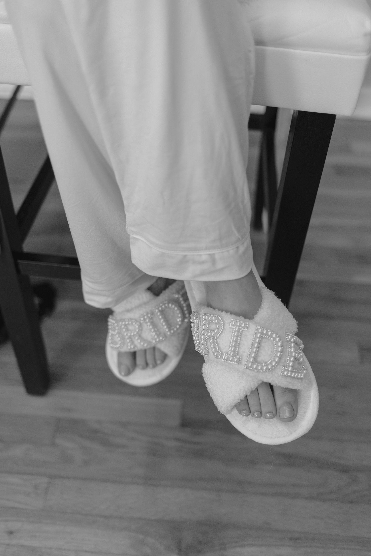 Black and white photo of bride wearing fluffy white BRIDE slippers while seated on a chair.
