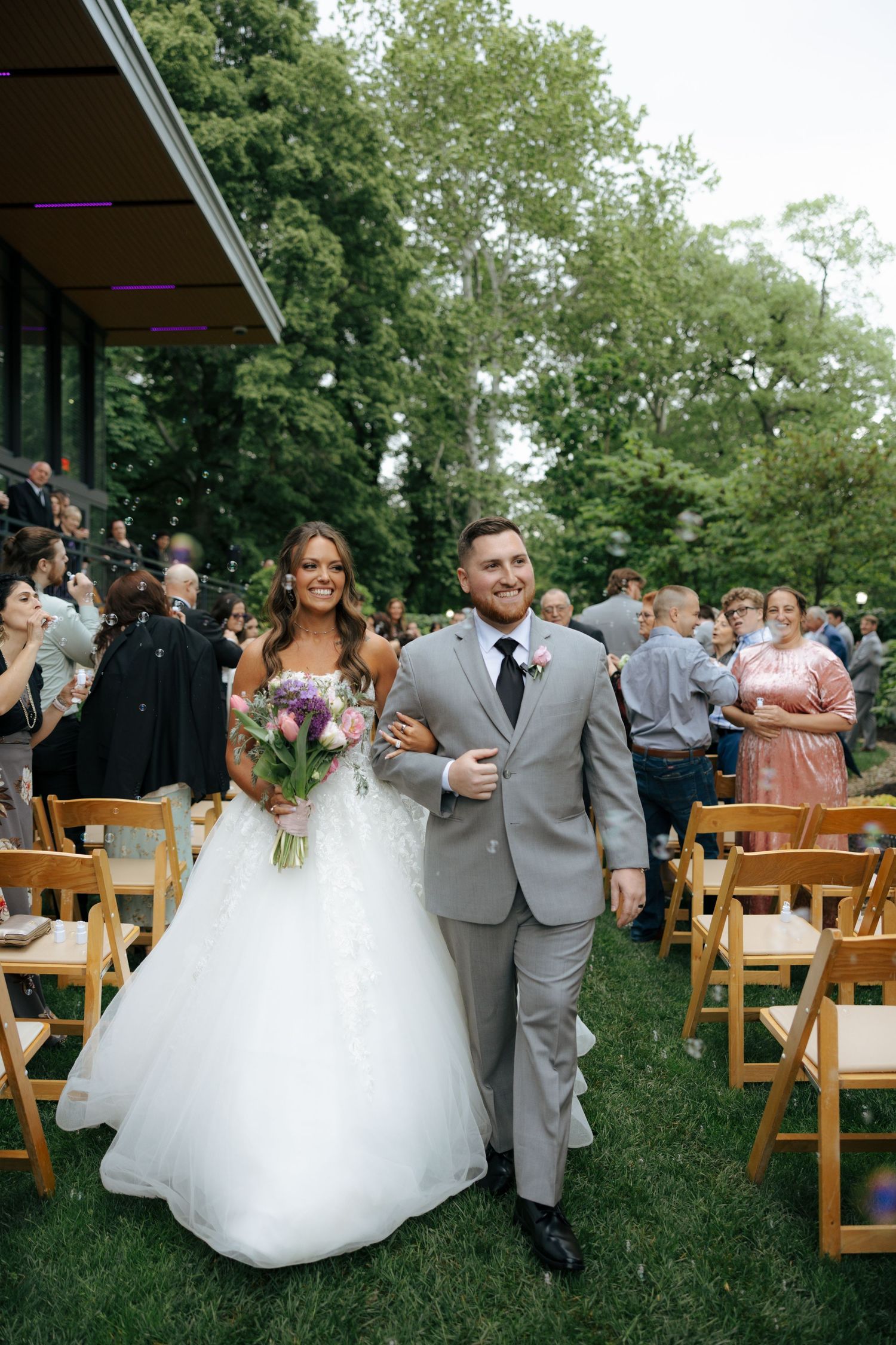 Smiling bride and groom walk down wedding aisle surrounded by seated guests at outdoor ceremony.