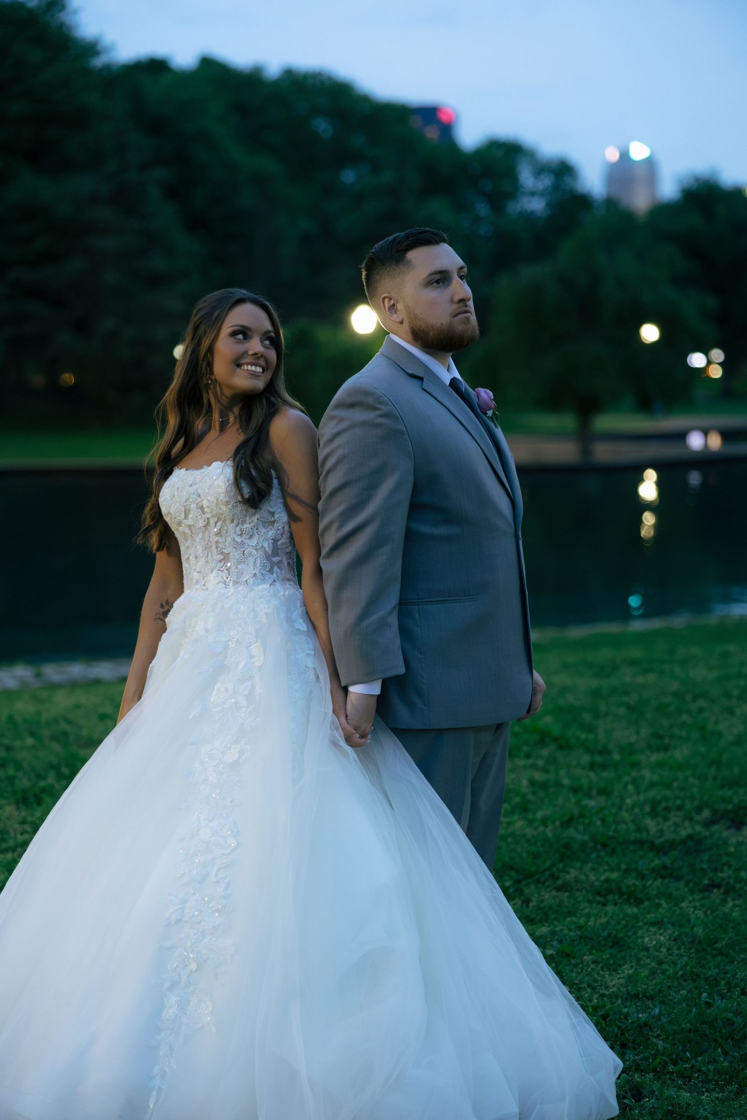 Smiling bride in lace ballgown and groom in gray suit pose back to back near a pond at twilight.