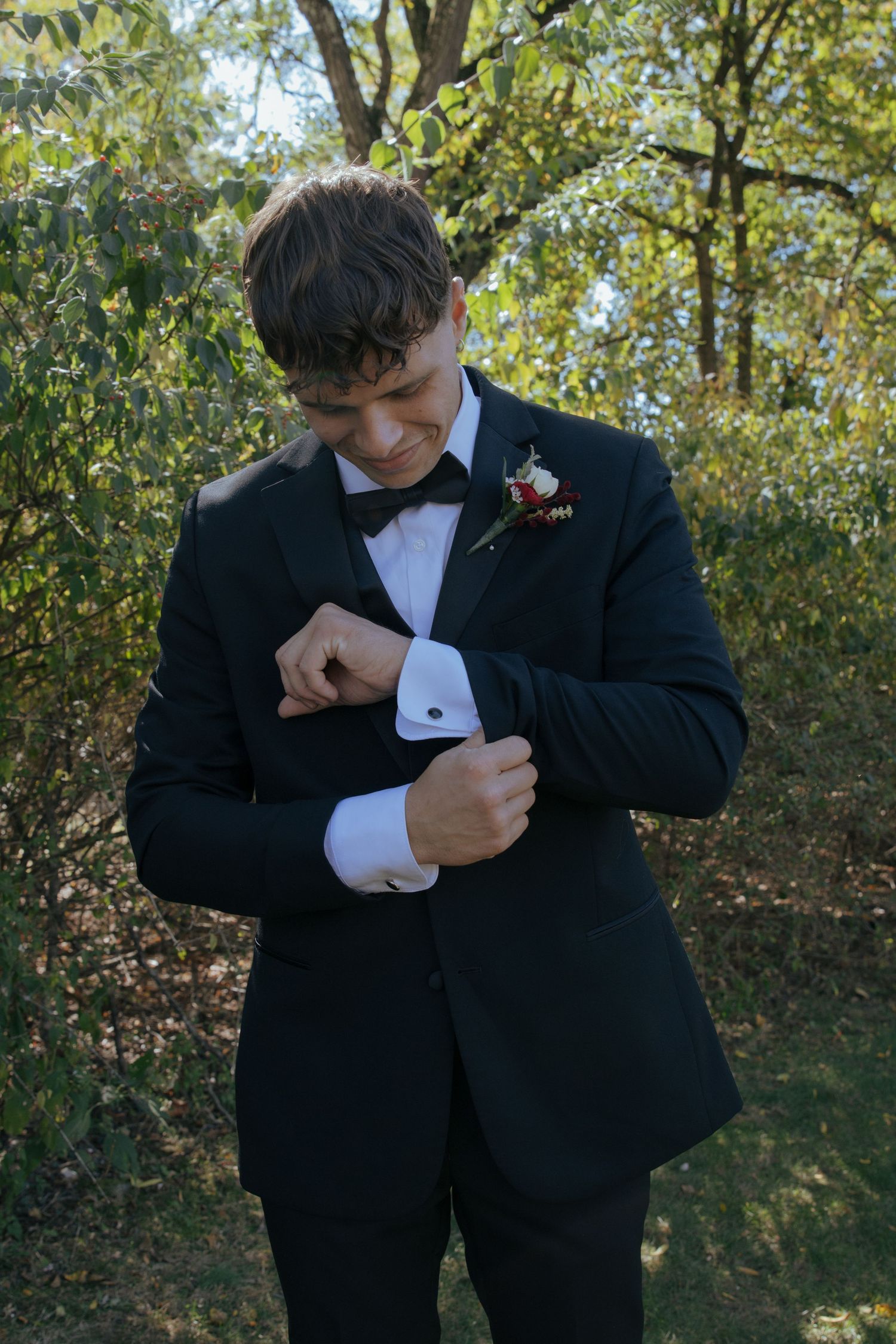 Groom in black tuxedo adjusting cufflinks with floral boutonniere outdoors.