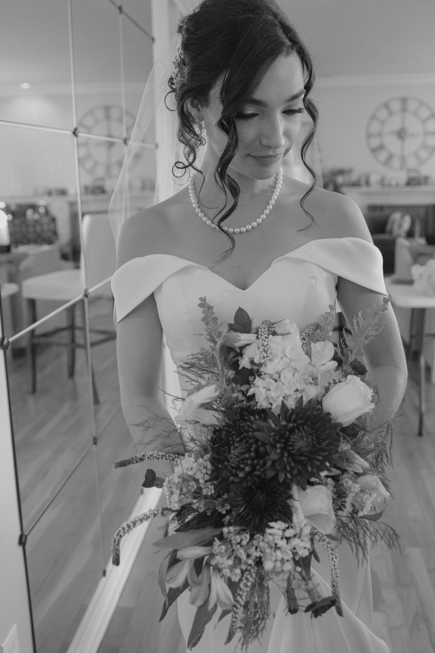 Bride in off-shoulder gown holding floral bouquet, wearing pearl necklace, black and white wedding photo.