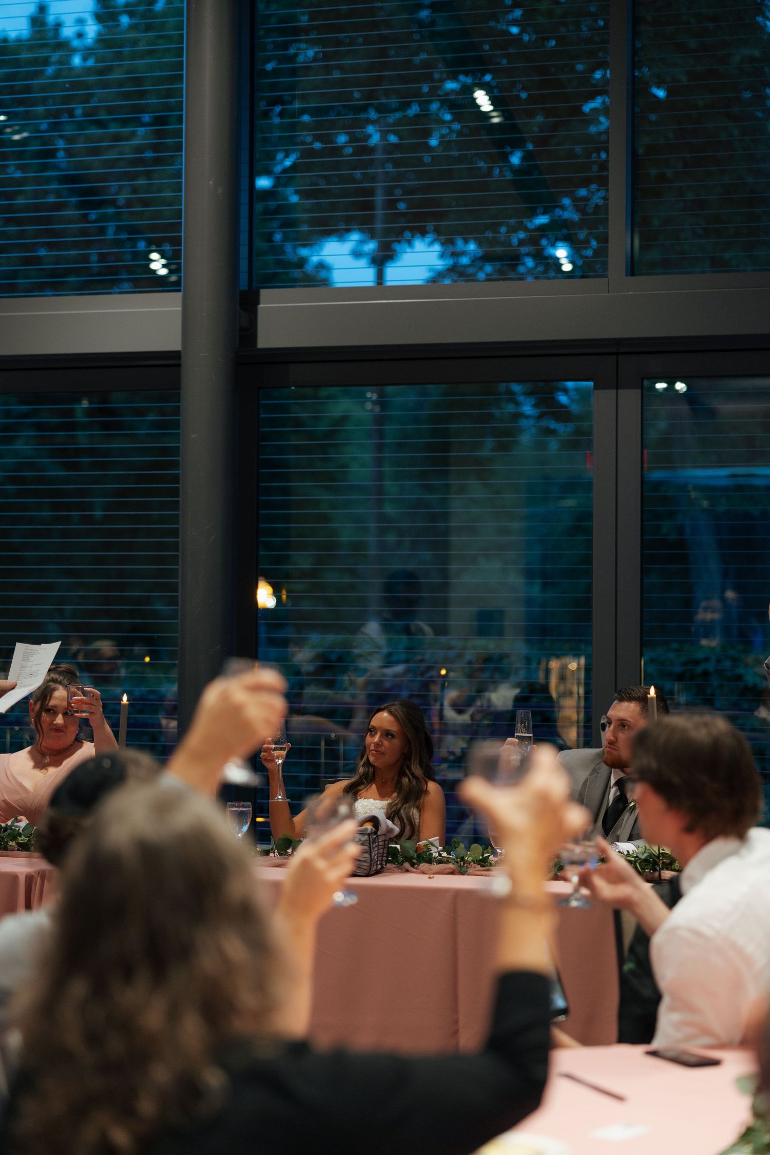 Wedding party raises glasses in toast to smiling bride at head table during evening reception celebration.