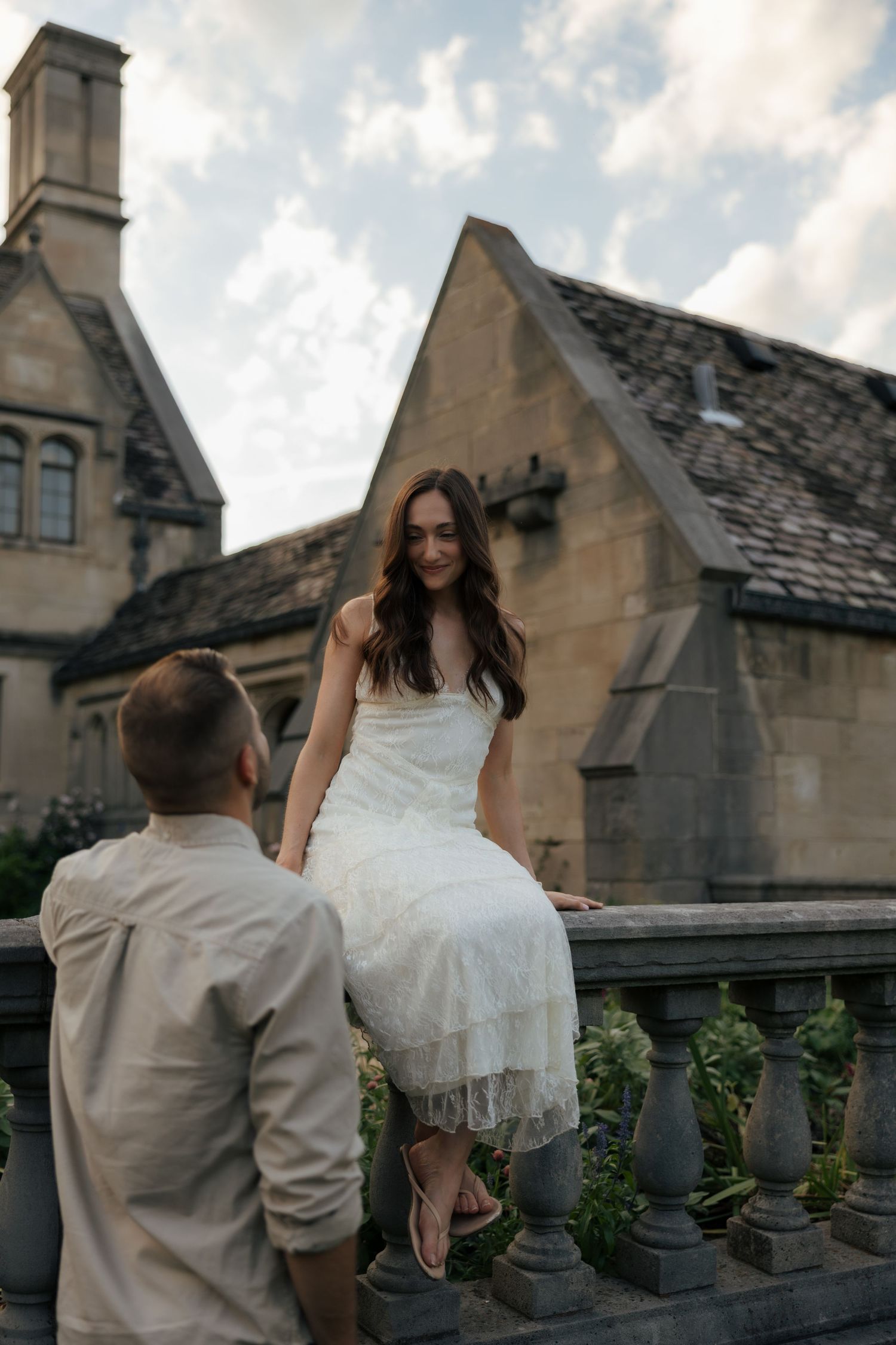 Bride in white lace dress sits on stone balustrade smiling at groom outside historic manor.
