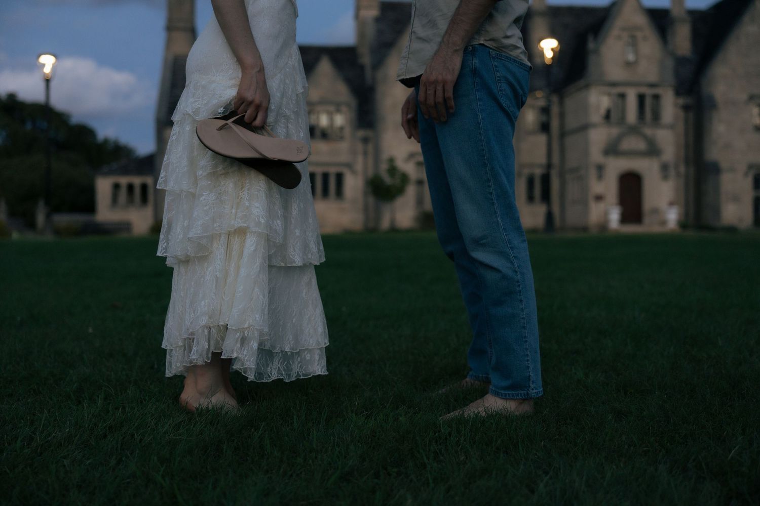 Barefoot bride holding shoes stands with groom on grass before grand historic manor at dusk.