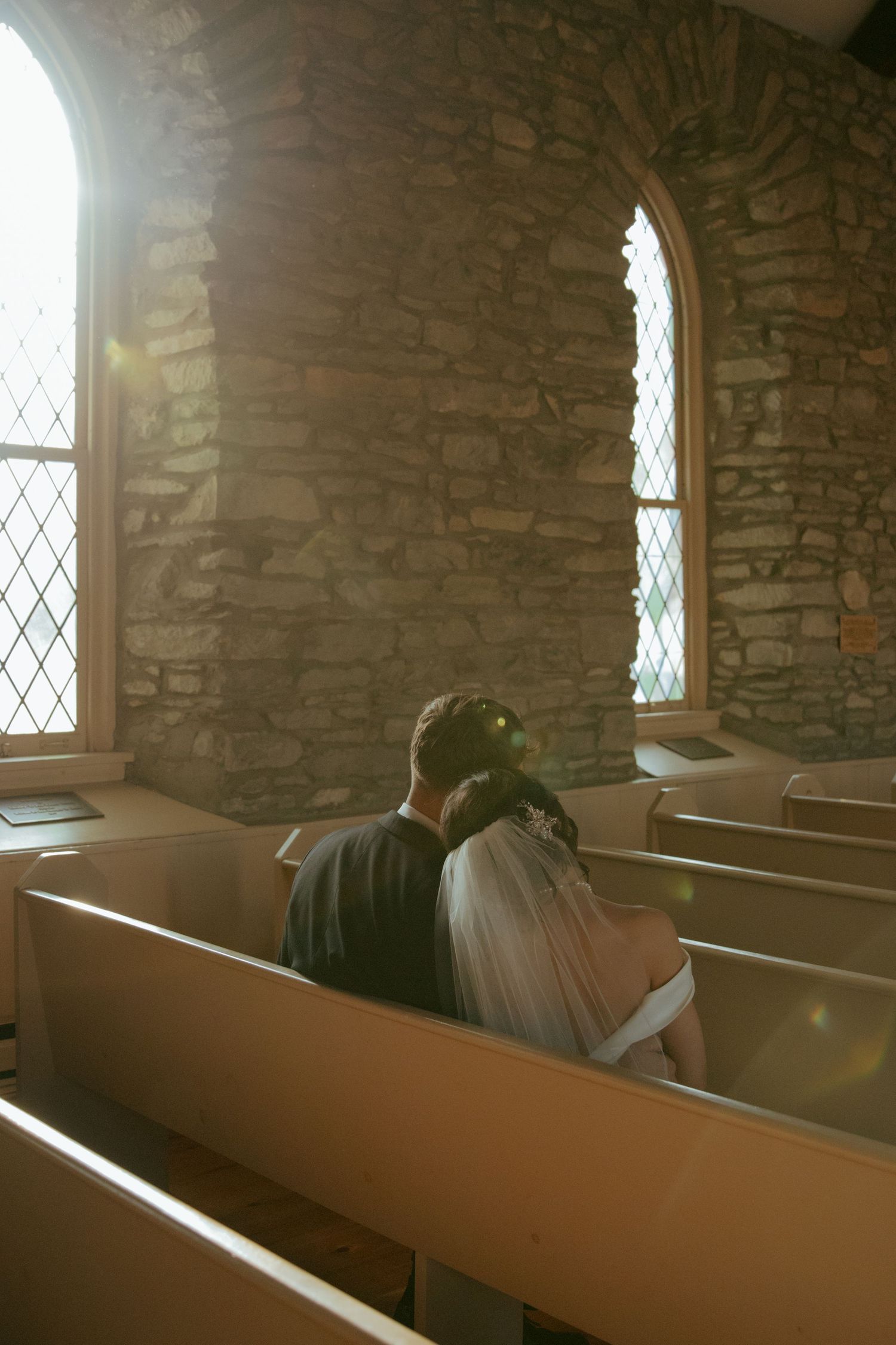 Sunlight streams through diamond-paned windows in a rustic stone church, illuminating wooden pews in warm golden light.