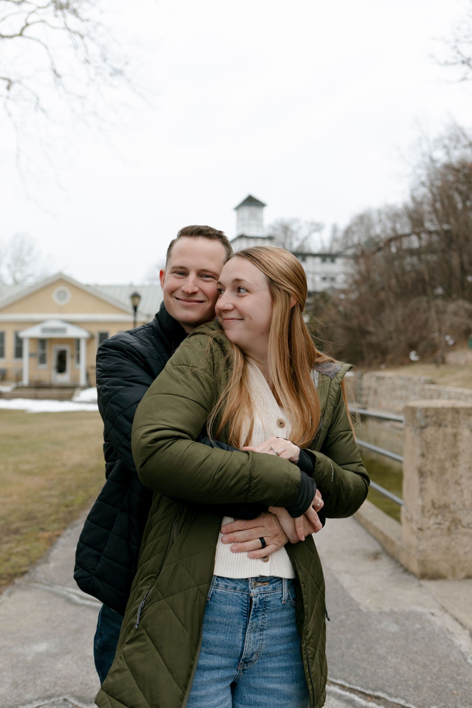 A couple embraces and laughs together on a path with a church building visible in the background.