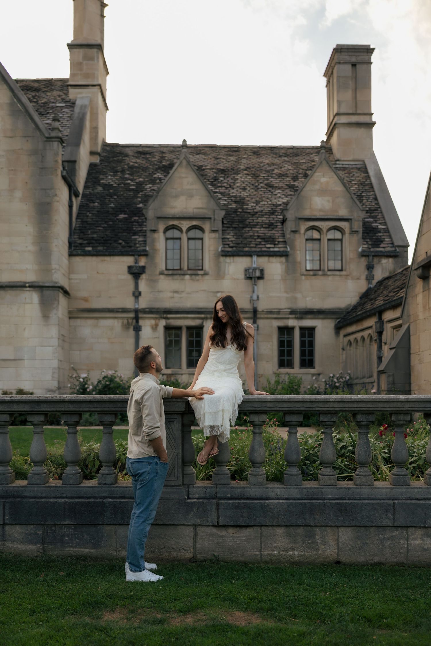 Couple sharing a romantic moment at a historic stone manor, woman in white dress sitting on balustrade.