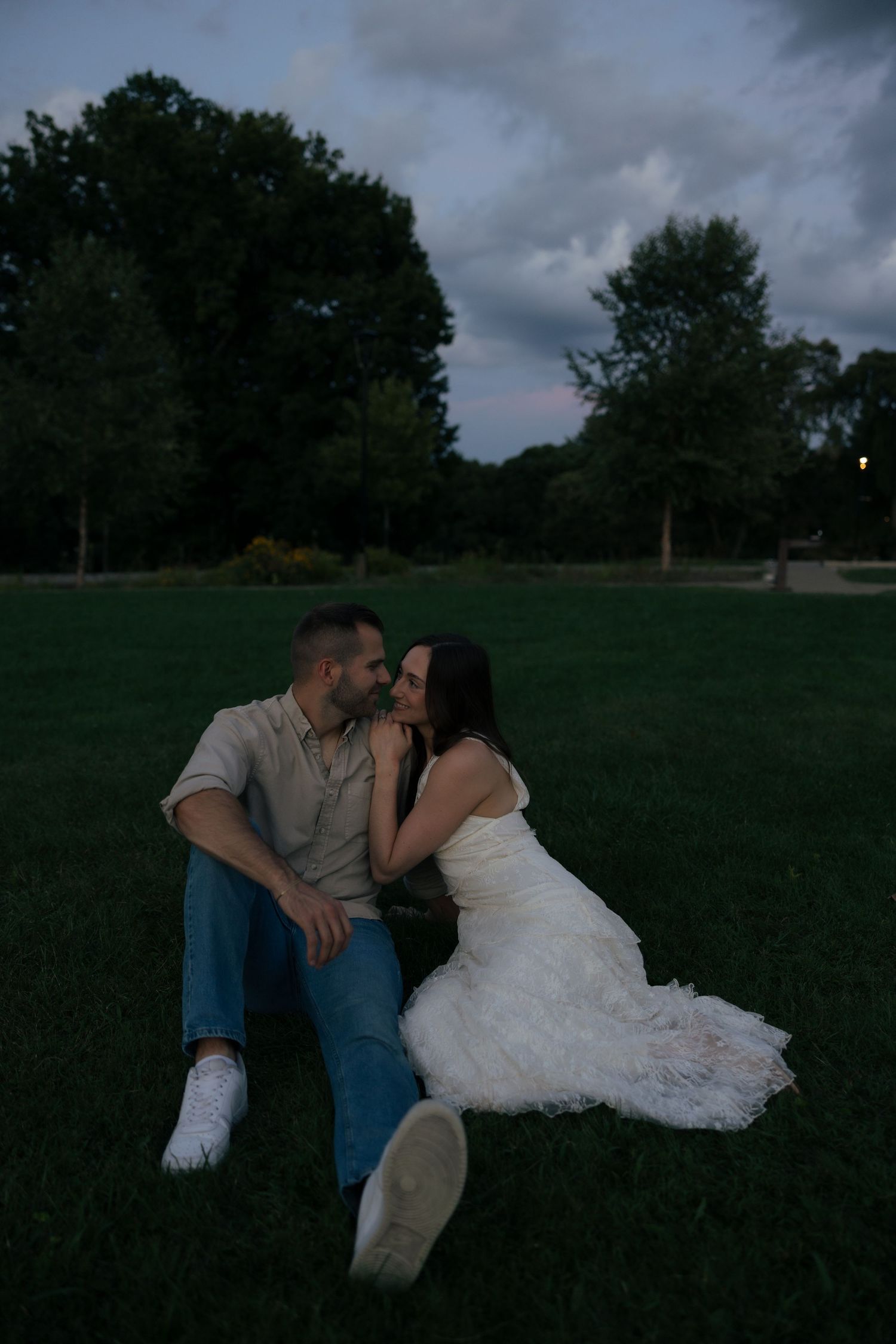 Couple sitting close on grass at dusk near trees, sharing an intimate moment.