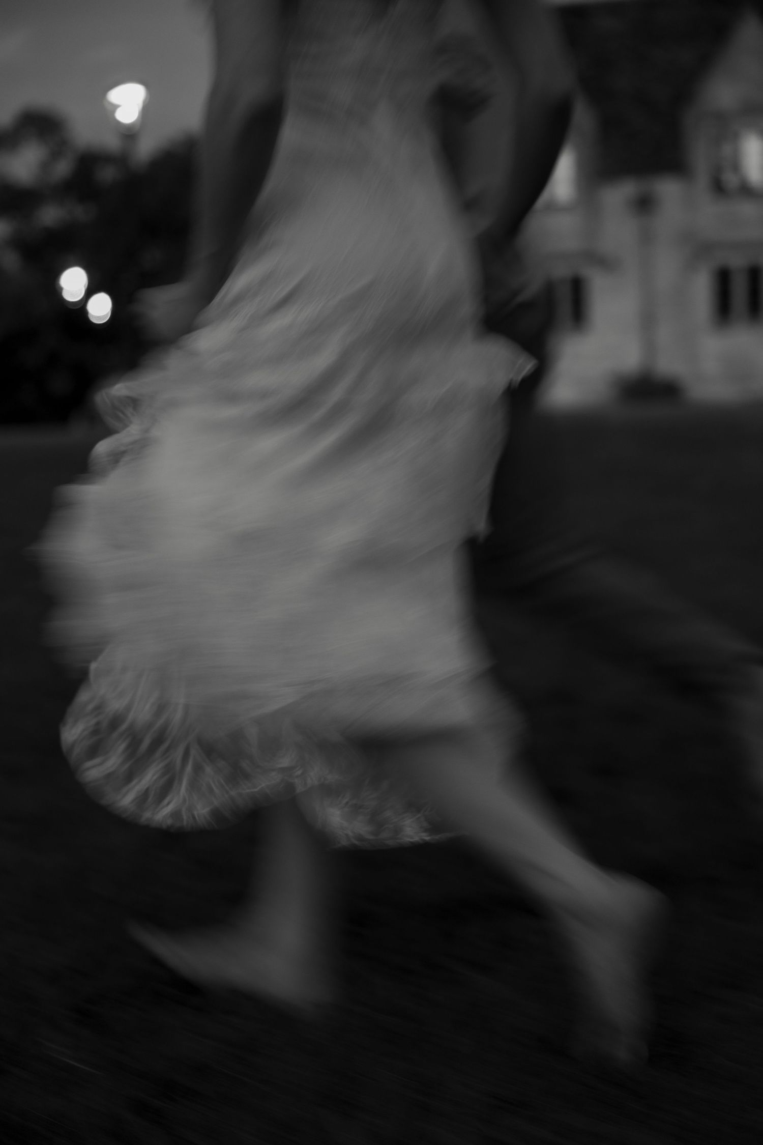 Blurred black and white motion shot of a woman in a flowing white dress running at dusk near a building.