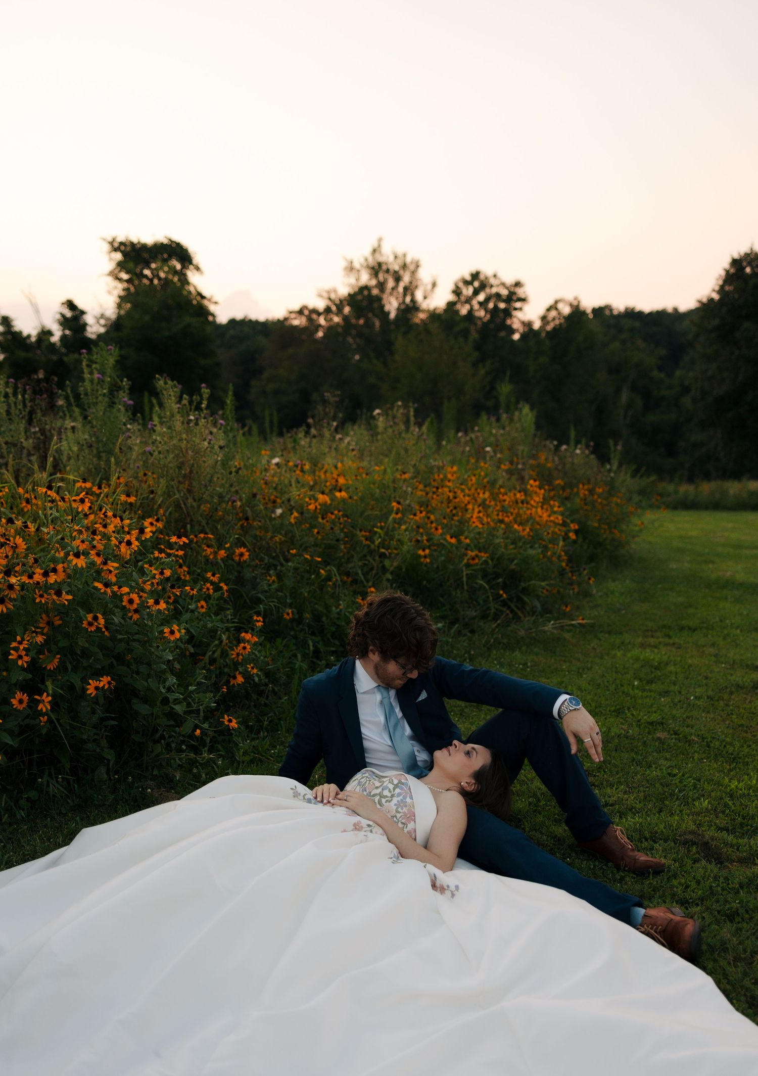 Couple in wedding attire recline together on grass beside vibrant orange wildflowers at dusk.