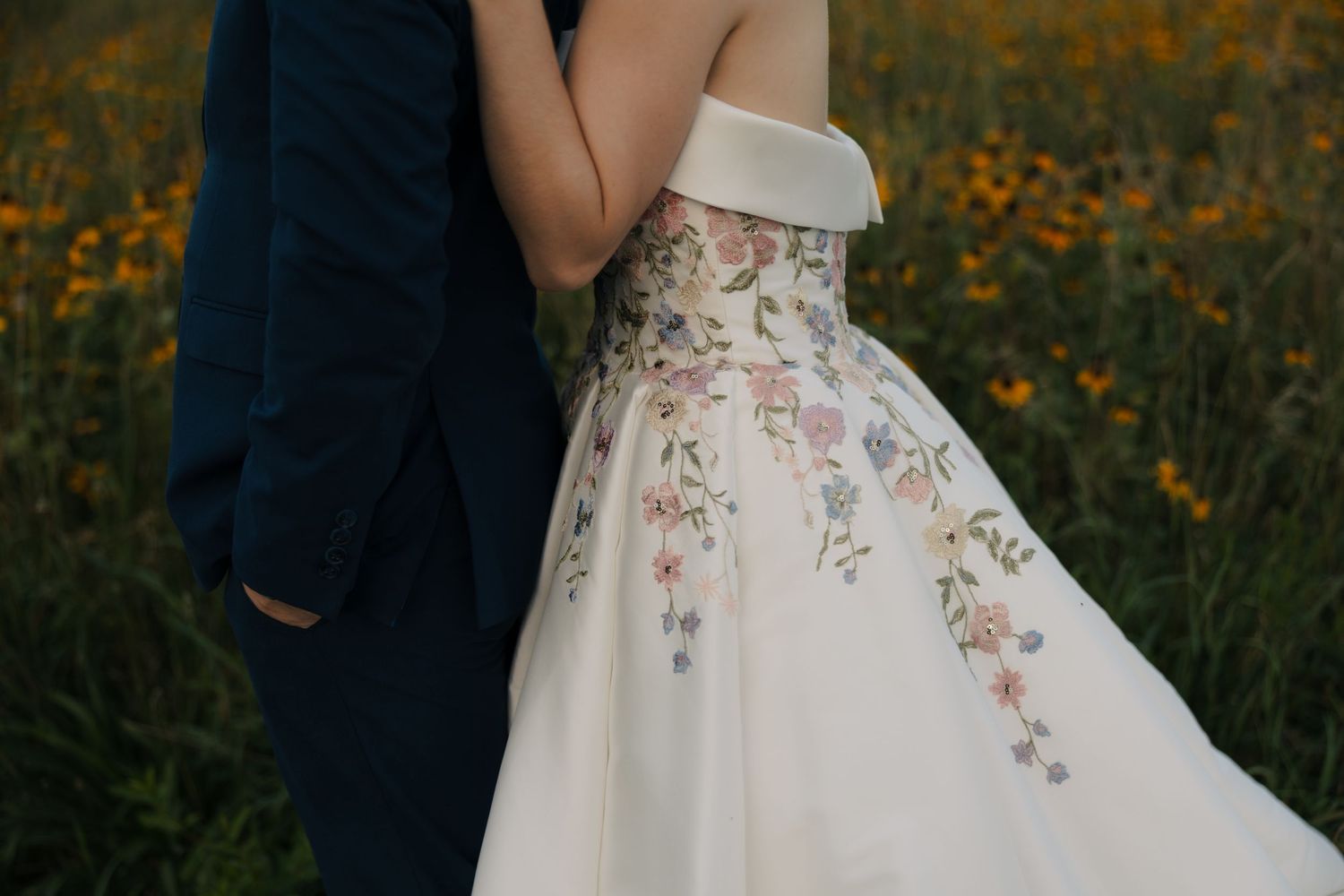 Close-up of bride in off-shoulder floral embroidered wedding dress standing with groom in yellow flower field.